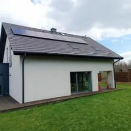 Nowoczesny dom jednorodzinny z panelami fotowoltaicznymi na brązowym dachu, widok z ogrodu z drewnianym tarasem i zielonym trawnikiem.