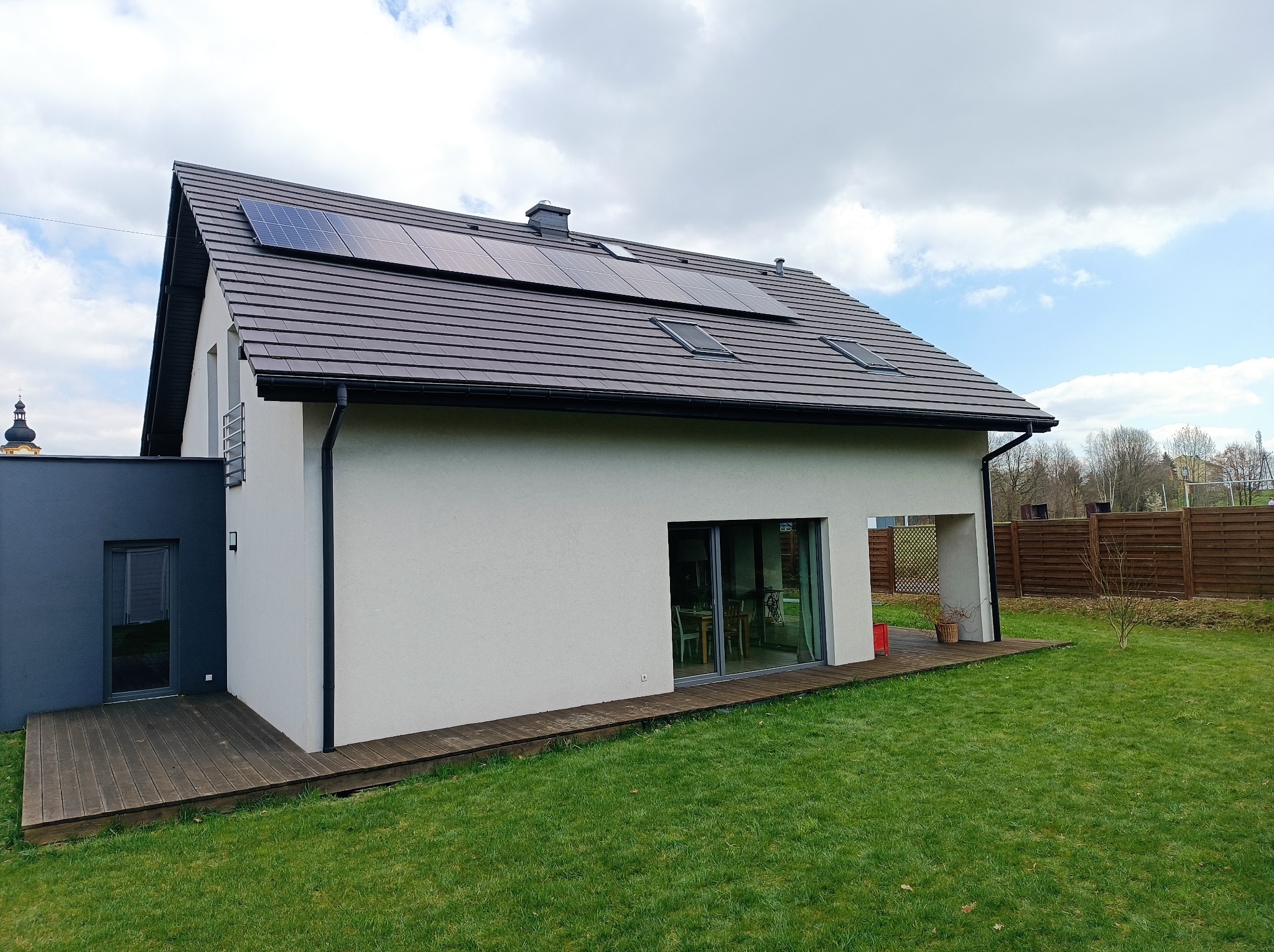 Nowoczesny dom jednorodzinny z panelami fotowoltaicznymi na brązowym dachu, widok z ogrodu z drewnianym tarasem i zielonym trawnikiem.