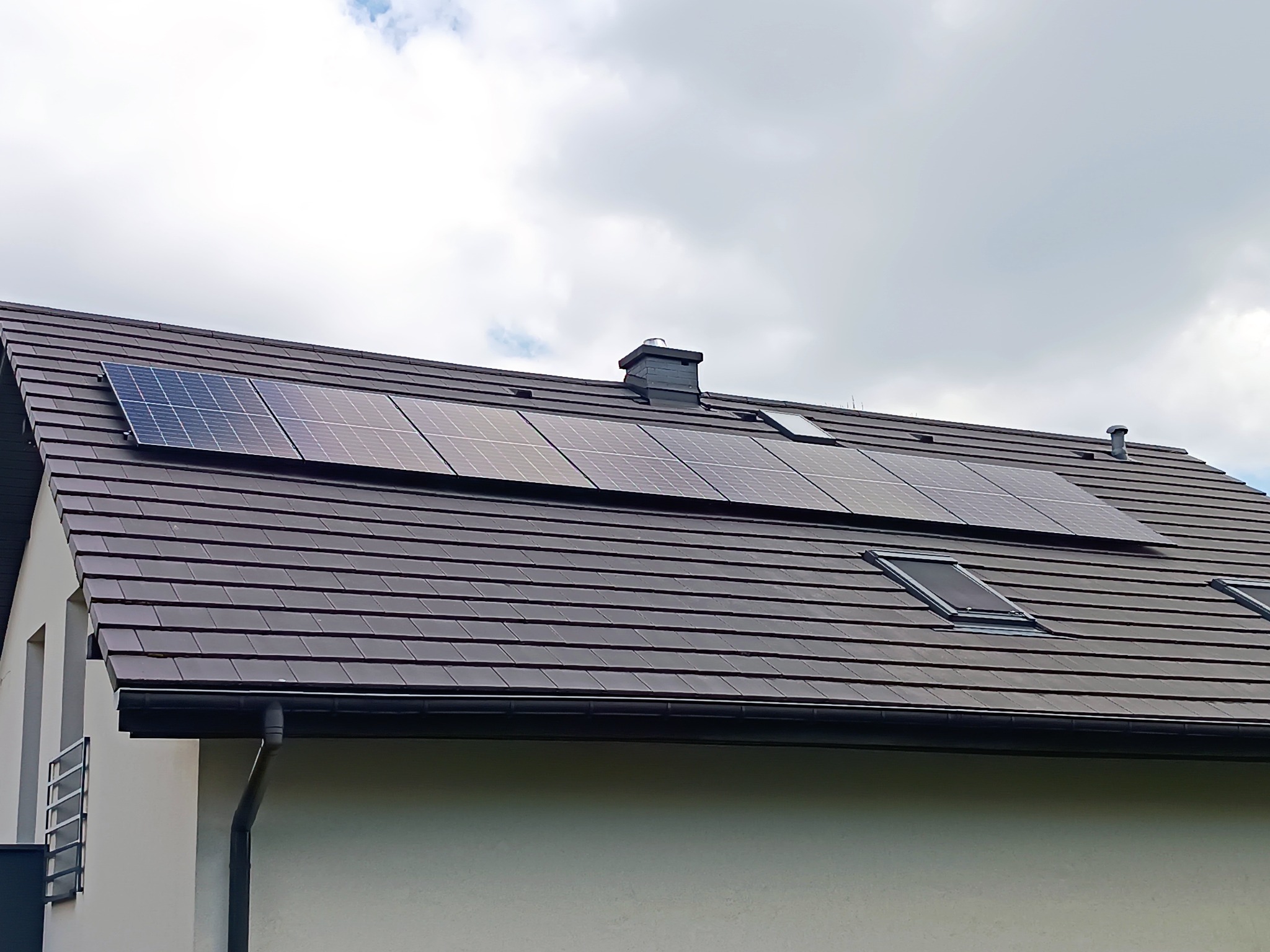 Panele słoneczne zamontowane na dachu domu z brązową dachówką, widoczne okna dachowe i komin, pochmurne niebo w tle.