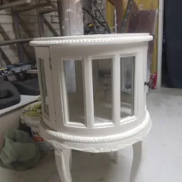 Okrągła, biała szafka z przeszklonymi drzwiczkami na giętych nóżkach, wykonana w Lęborku, stoi w warsztacie na tle ściany z cegły i drewnianych elementów.