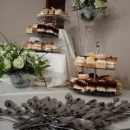 Trzypoziomowy tort ze słodkościami, ciastami i pączkami, dekorowany owocami, ustawiony na białym stole z dekoracjami kwiatowymi i stertą srebrnych łyżeczek.