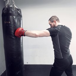 Trener personalny Michał - Gniezno - Mężczyzna w czarnej koszulce uderza w czarny worek treningowy w czerwonej rękawicy bokserskiej, widoczny monitoring na suficie.