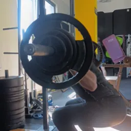 Osoba wykonuje przysiad ze sztangą w siłowni, na tle żółtej kolumny i okna. Widoczny sprzęt do ćwiczeń i podest.