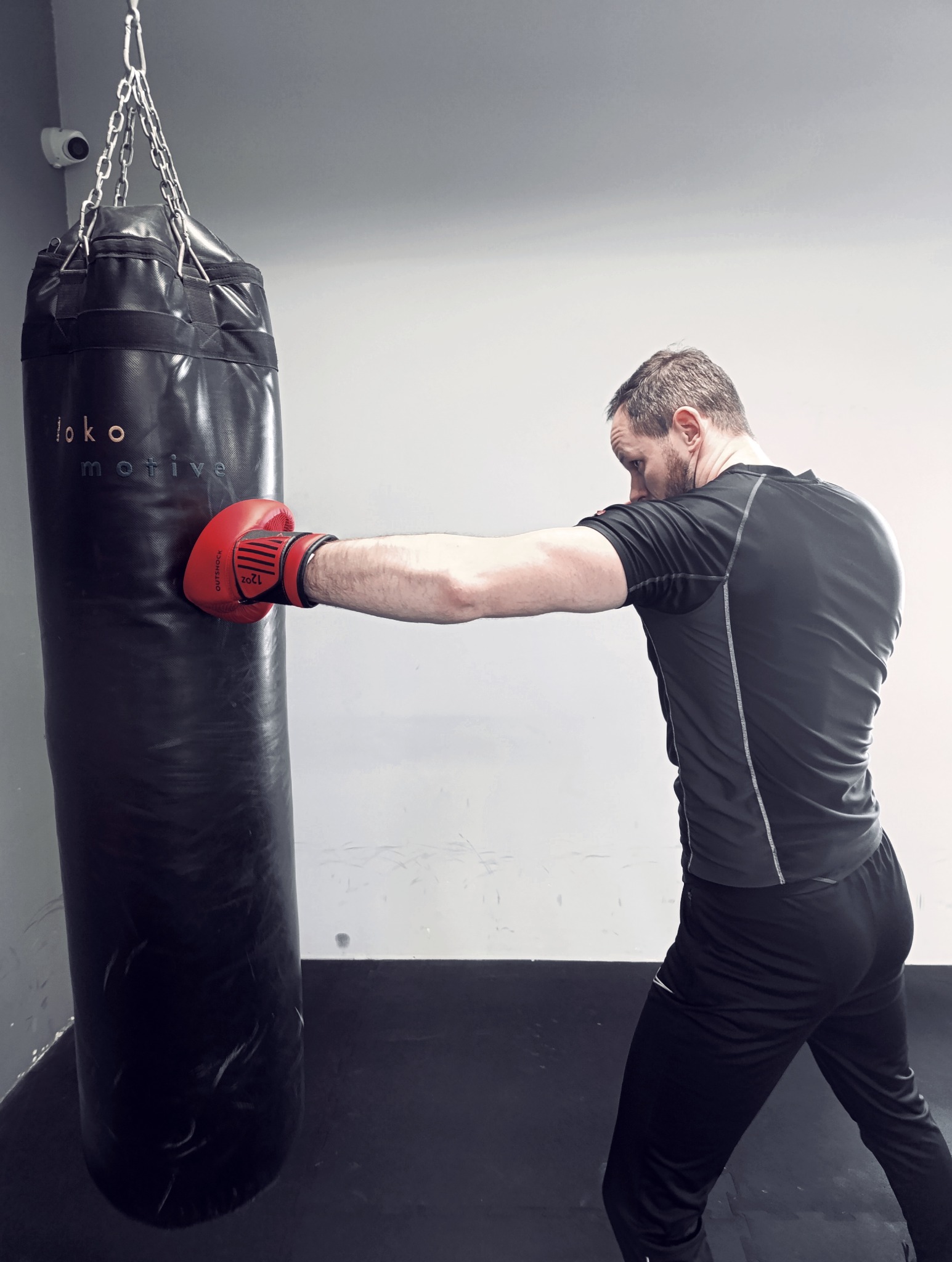 Mężczyzna w czarnej koszulce uderza w czarny worek treningowy w czerwonej rękawicy bokserskiej, widoczny monitoring na suficie.