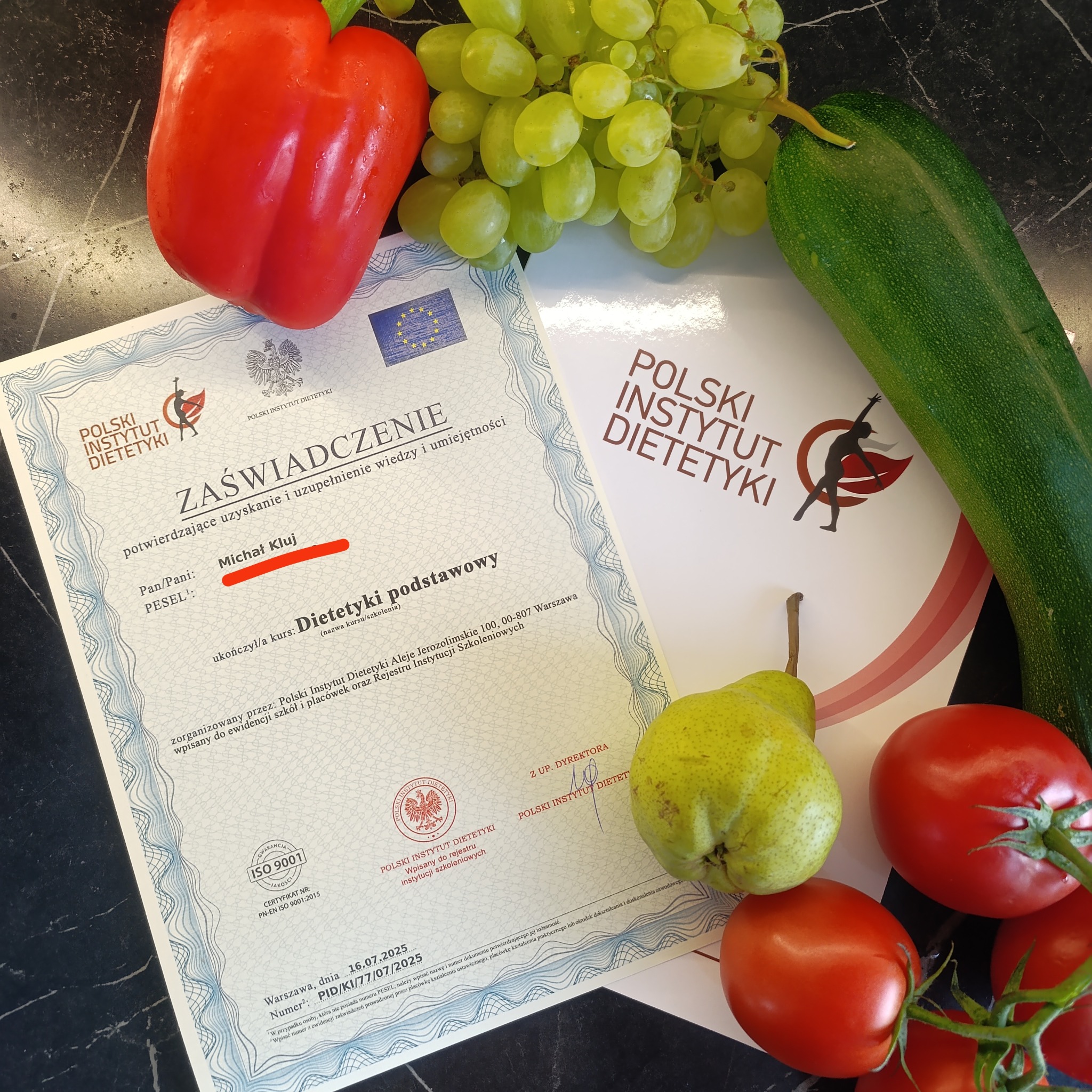 Certyfikat dietetyka Michała Kluj z warzywami i owocami na blacie: papryka, winogrona, cukinia, gruszka, pomidory. Ukończony kurs dietetyki podstawowej.