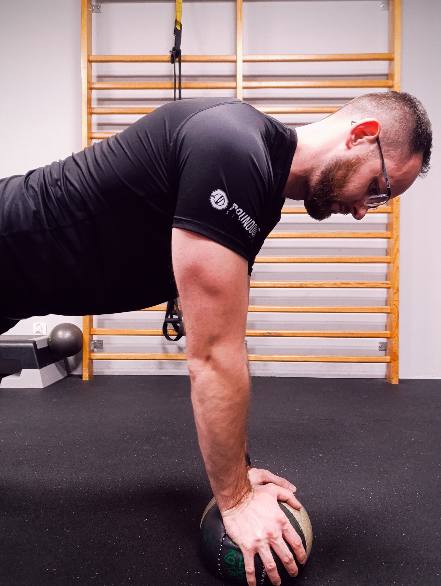 Mężczyzna w czarnej koszulce wykonuje pompkę na piłce lekarskiej w siłowni. W tle drabinki gimnastyczne i step. Trening siłowy z obciążeniem.