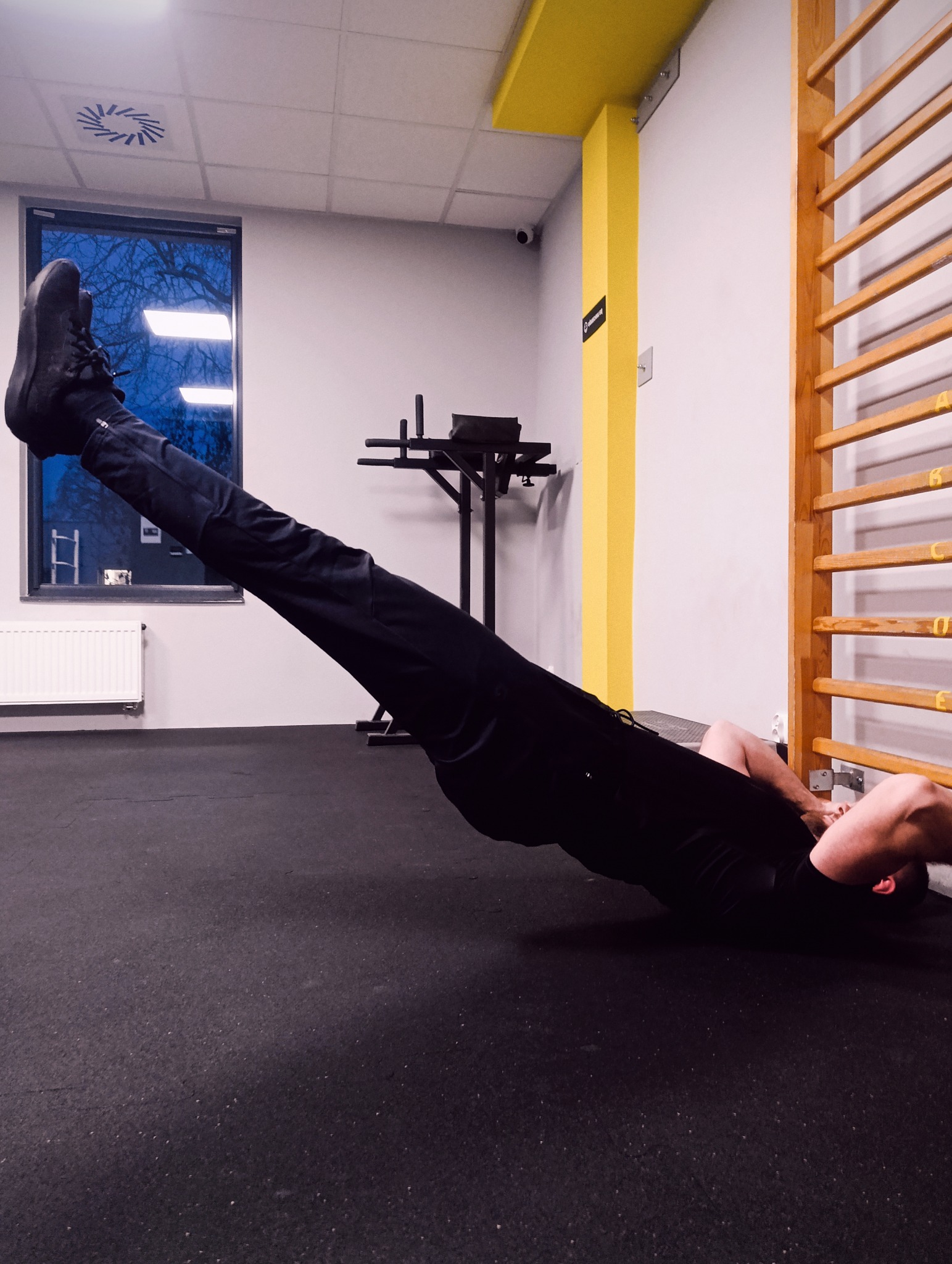 Osoba w czarnym stroju wykonuje ćwiczenie unoszenia bioder w sali gimnastycznej z drabinkami i oknem z widokiem na nocne niebo.