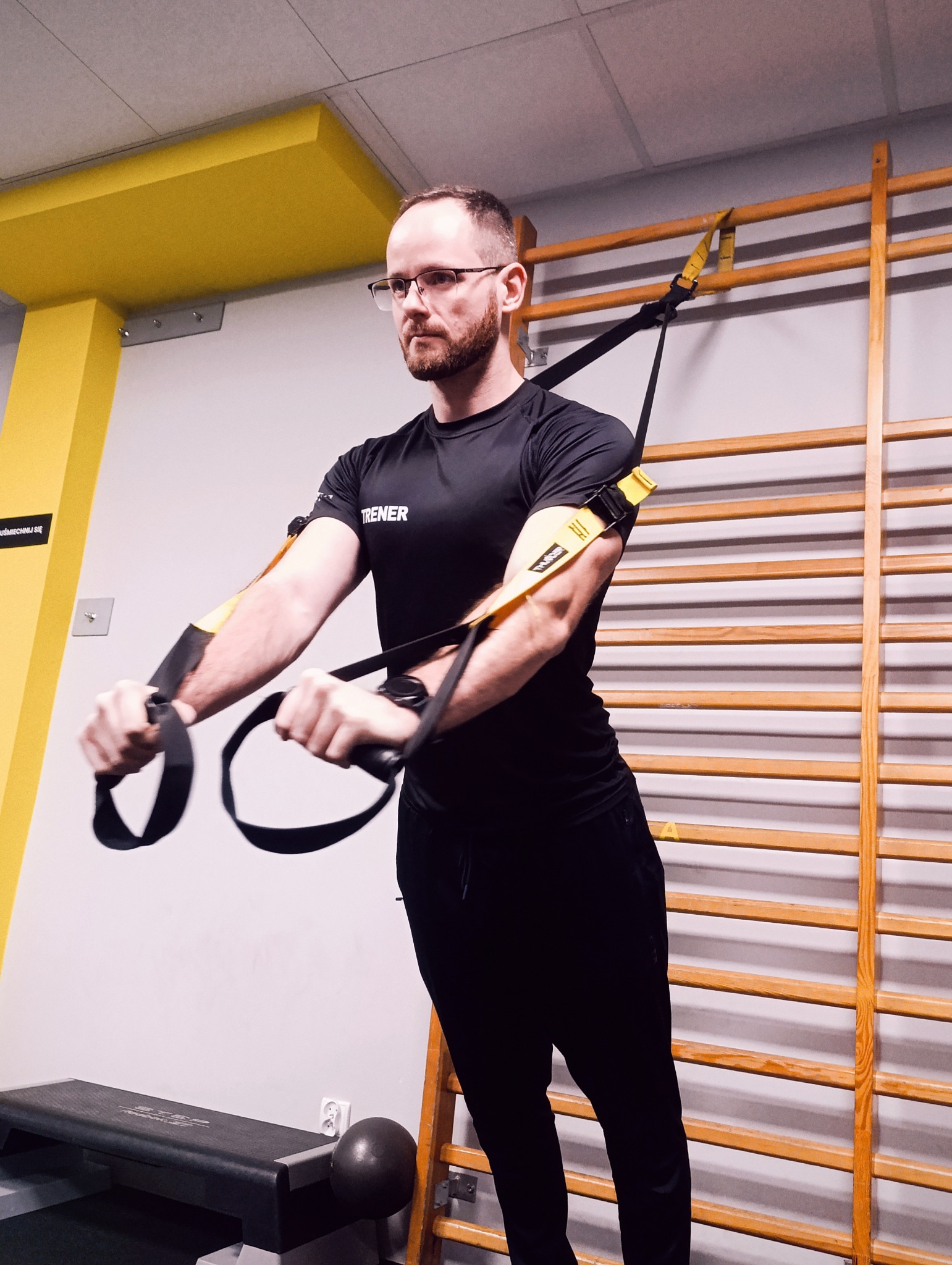 Mężczyzna w czarnym stroju sportowym ćwiczy na TRX w siłowni. W tle drabinki gimnastyczne i żółty element konstrukcyjny. Widoczny napis 'TRENER' na koszulce.