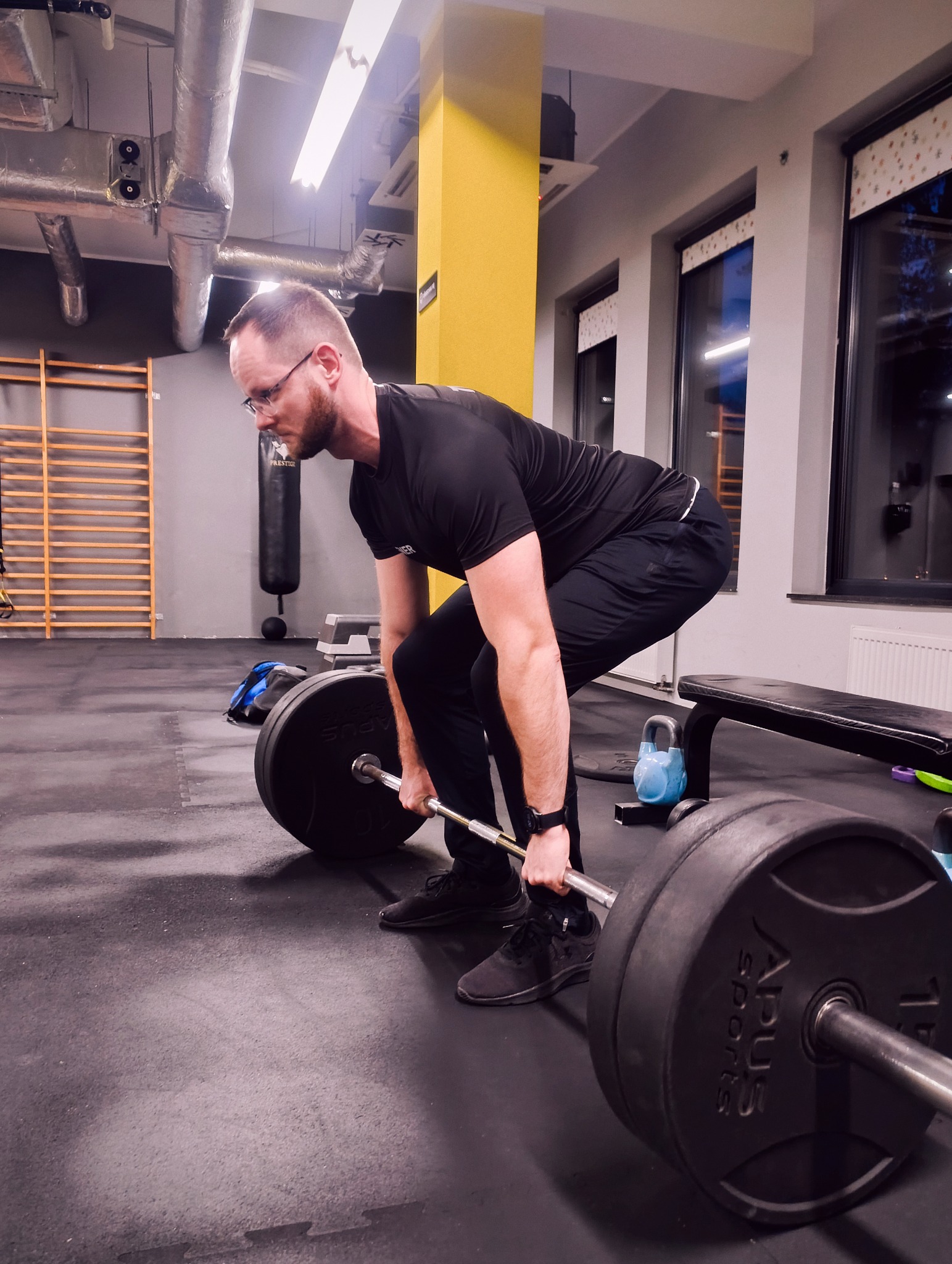 Mężczyzna w czarnym stroju sportowym wykonuje martwy ciąg ze sztangą w siłowni. Widoczne obciążenia, podłoga z mat, drabinki i okna w tle.