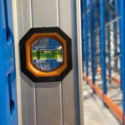 Poziomnica przyłożona do pionowej belki regału magazynowego, w tle widoczne niebieskie ramy regałowe.