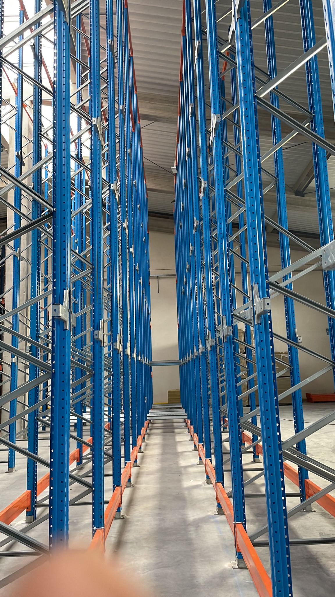 Długi rząd niebieskich, metalowych regałów magazynowych z pomarańczowymi poprzeczkami w jasnym magazynie, widok z perspektywy podłogi, z lekkim rozmyciem u dołu kadru.
