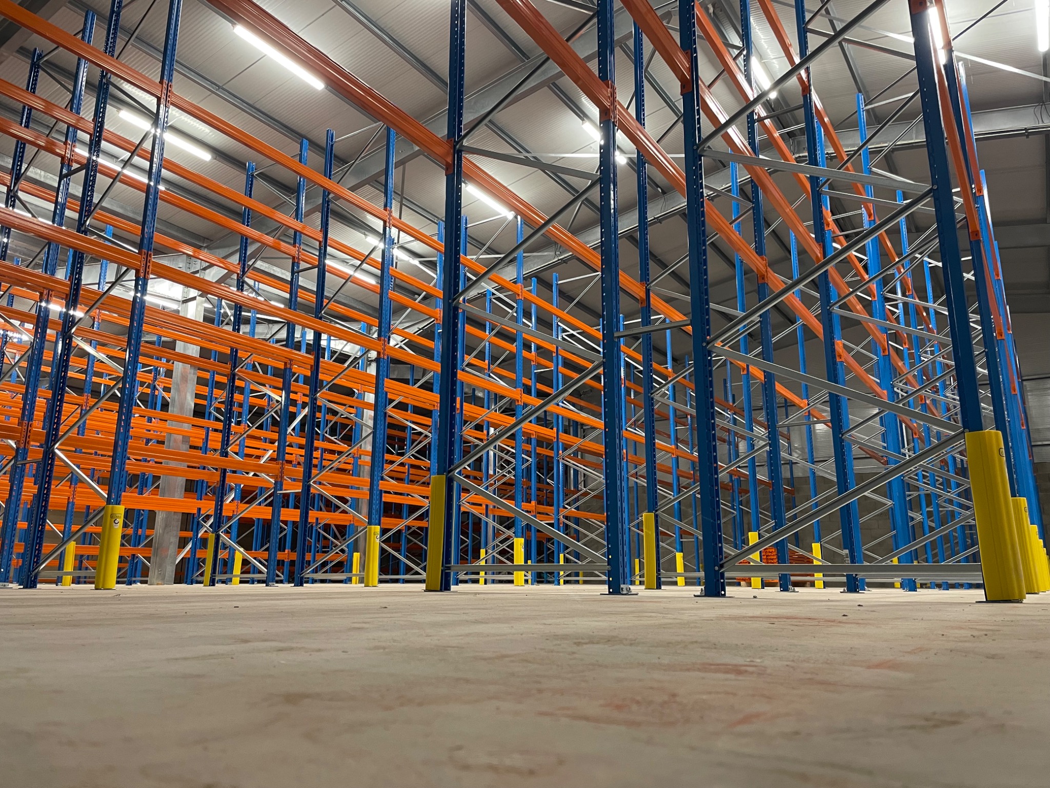 Nowe, niebiesko-pomarańczowe regały magazynowe z żółtymi ochraniaczami na słupach, sfotografowane z niskiej perspektywy, ukazujące przestronne wnętrze magazynu z oświetleniem sufitowym.