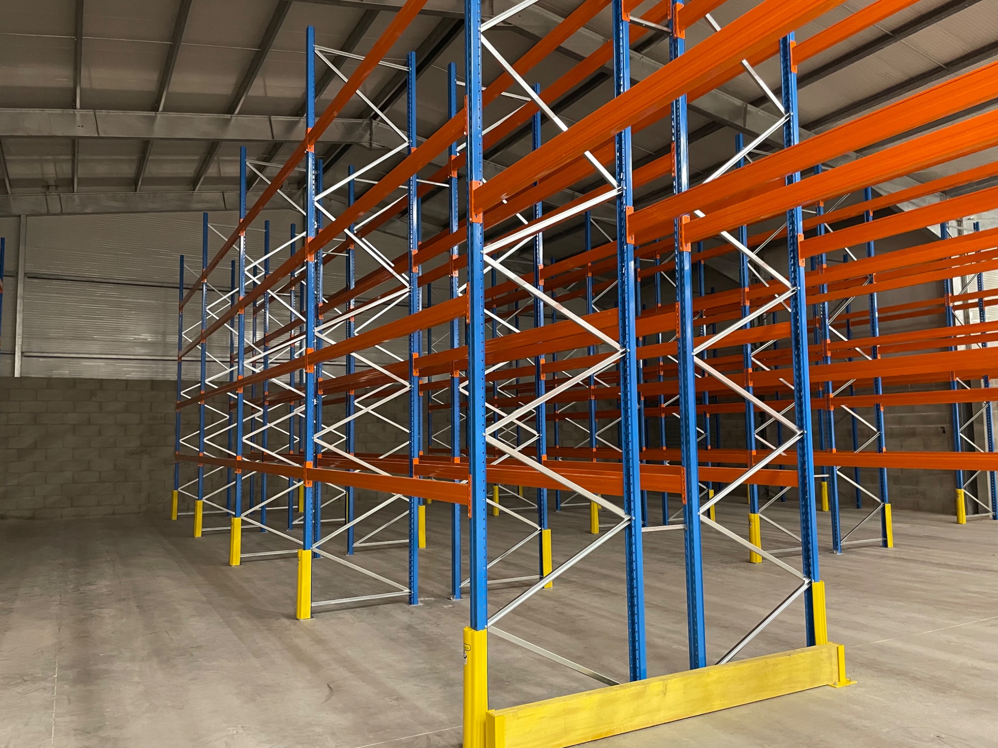 Puste, nowe regały magazynowe z niebieskimi słupkami, pomarańczowymi poprzeczkami i żółtymi osłonami zabezpieczającymi, ustawione w rzędach w hali magazynowej.
