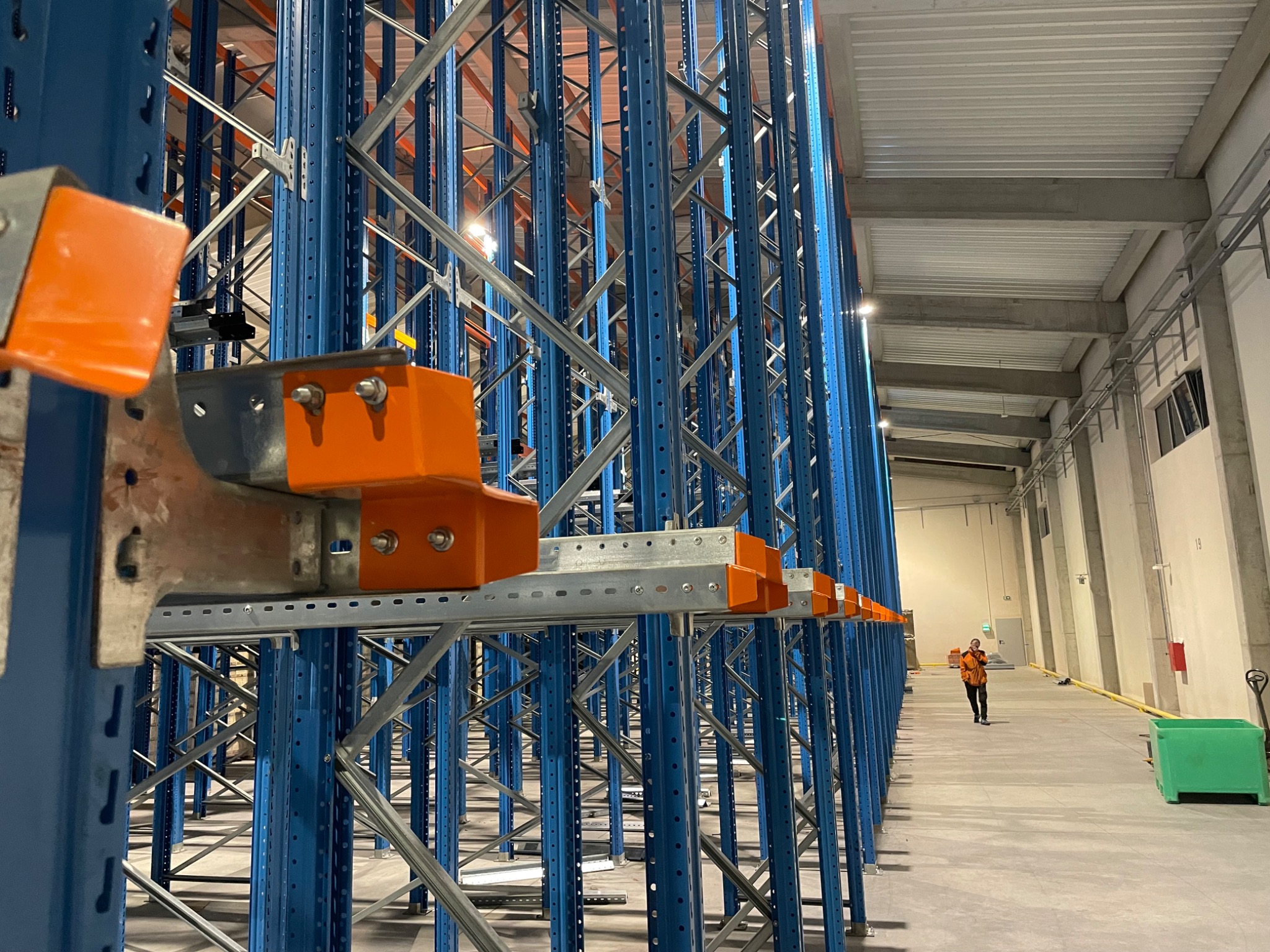 Wysokie, niebieskie regały magazynowe z pomarańczowymi elementami mocującymi, widoczne z perspektywy niskiego kąta w przestronnym magazynie z pracownikiem w oddali.