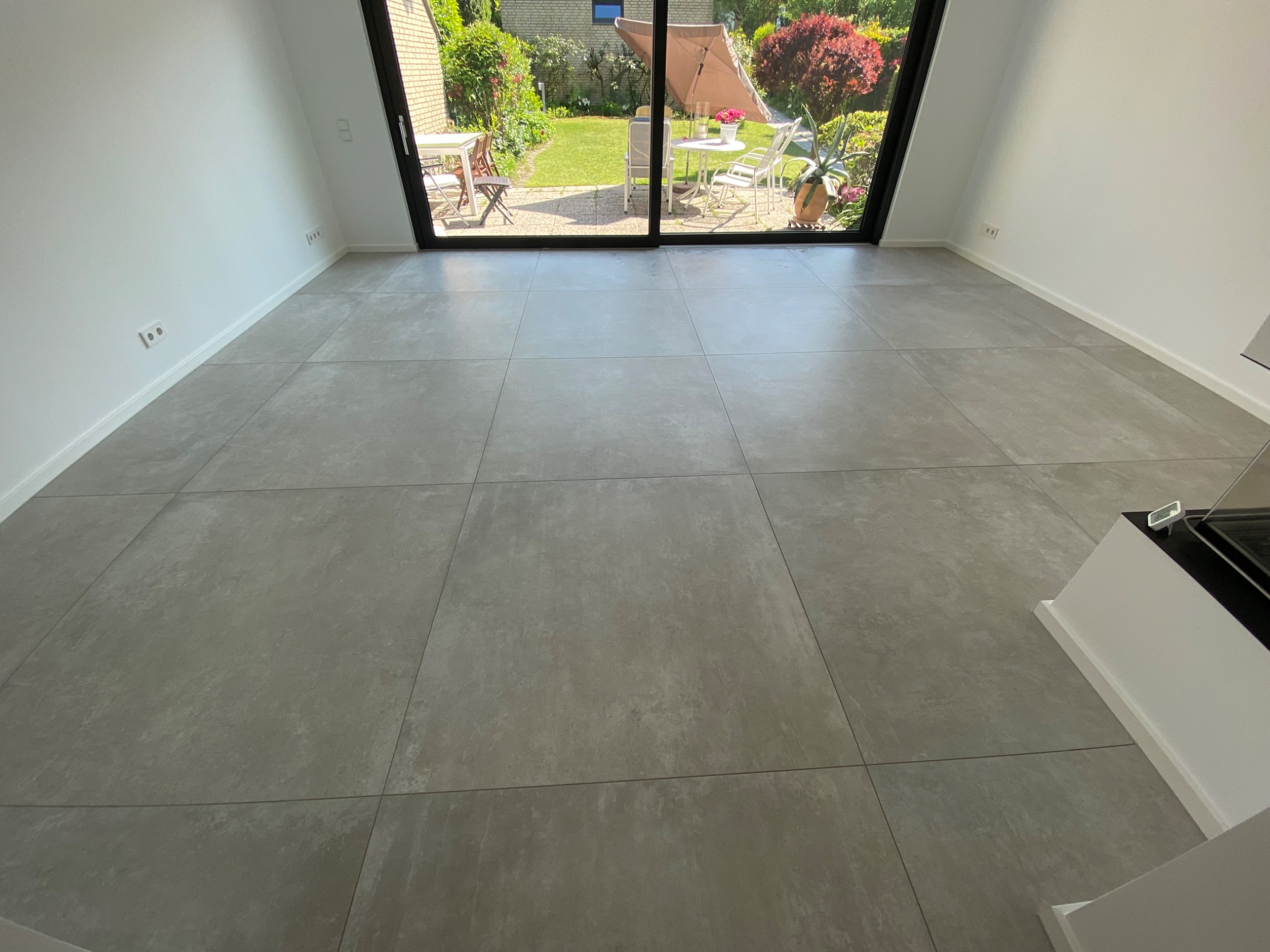 Nowoczesna podłoga z szarych, matowych płytek w przestronnym salonie z widokiem na zielony ogród przez duże, przesuwne drzwi tarasowe.