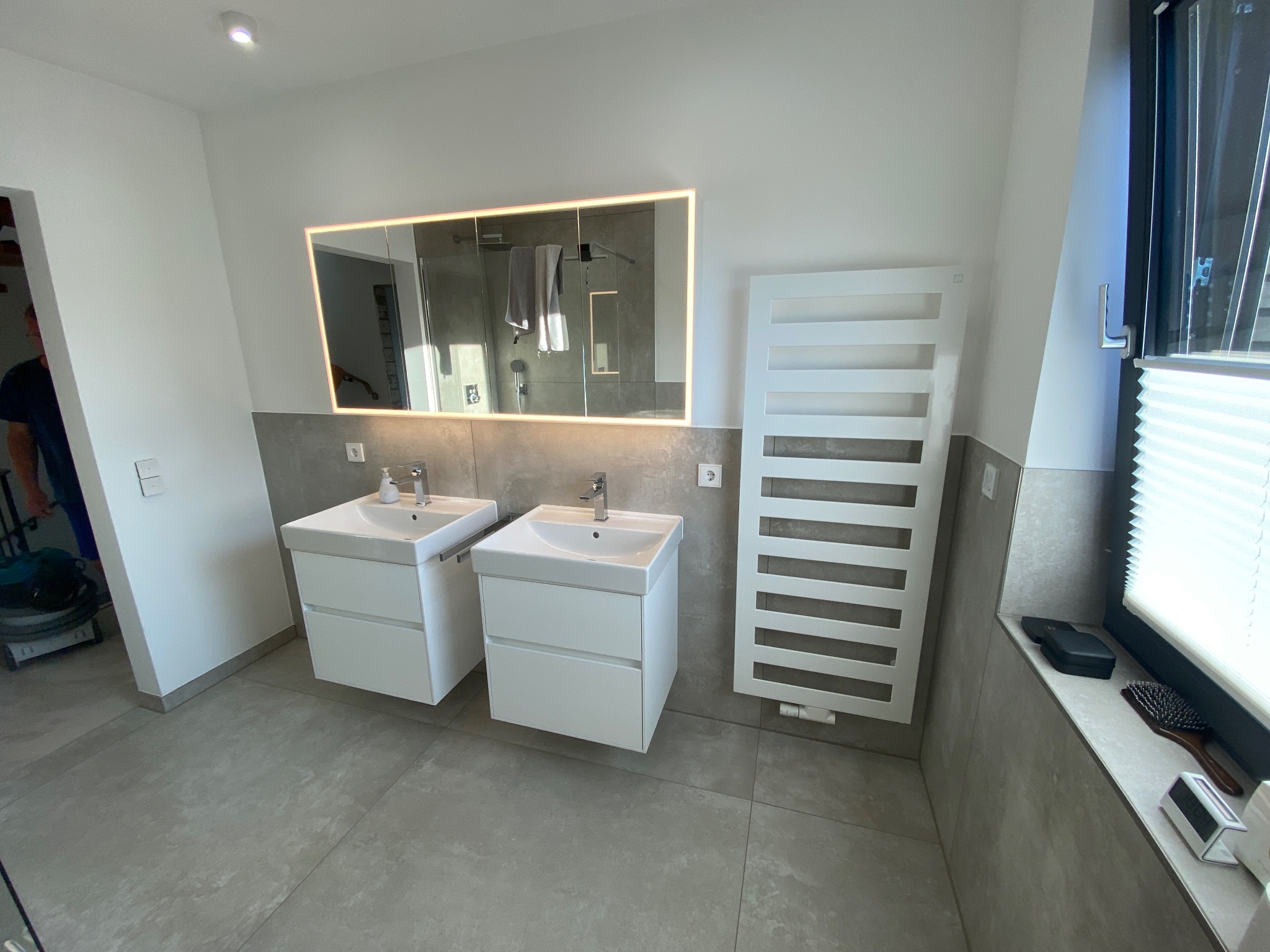 Nowoczesna łazienka z dwiema białymi umywalkami nablatowymi, podświetlanym lustrem, grzejnikiem drabinkowym i szarymi płytkami podłogowymi oraz ściennymi.