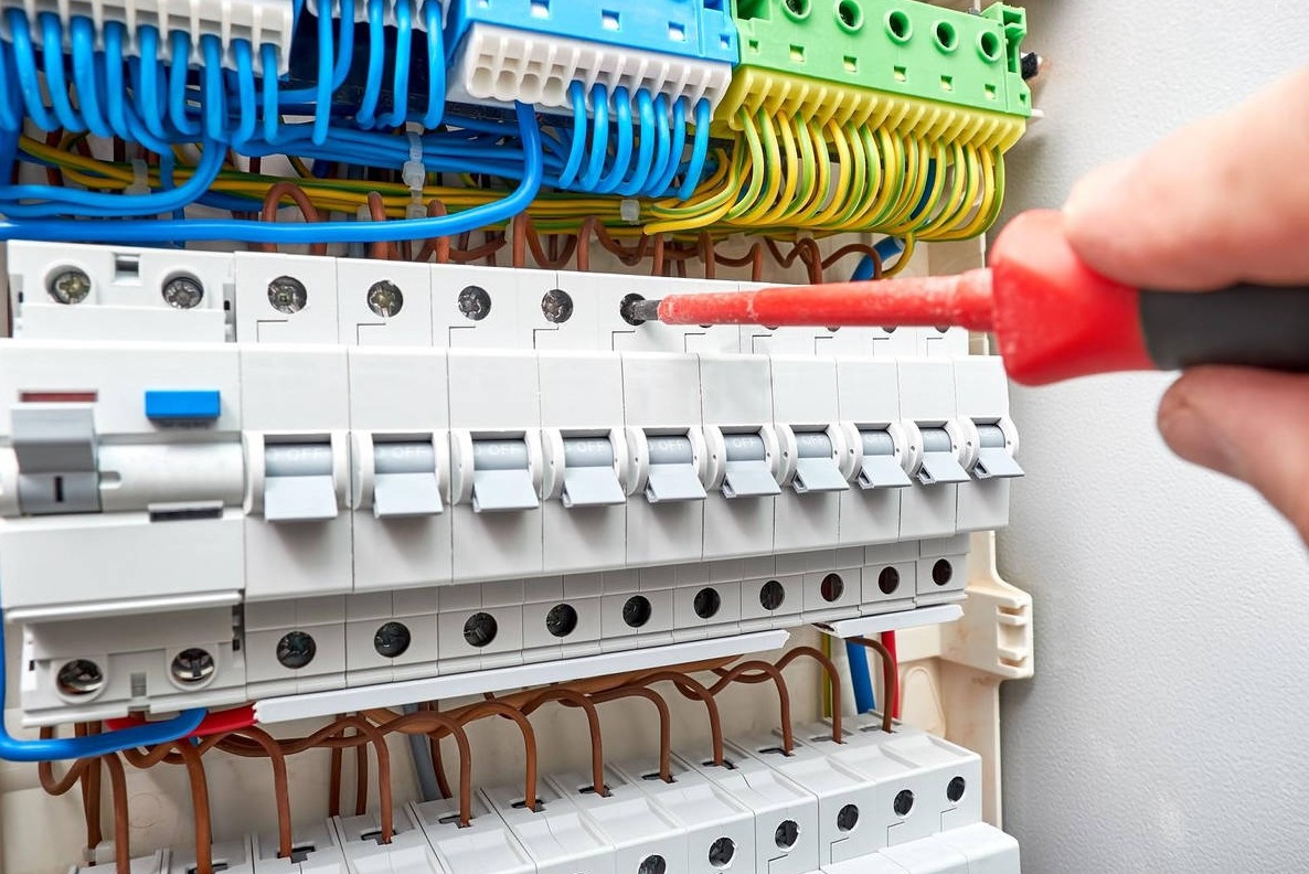 Elektryk dokręca śrubę w rozdzielnicy elektrycznej z bezpiecznikami, widoczne kolorowe przewody i czerwony śrubokręt.