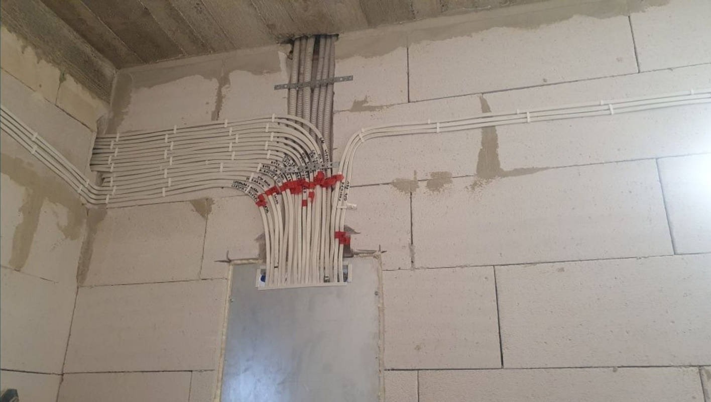 Białe przewody elektryczne ułożone w ścianie z betonu komórkowego, zabezpieczone czerwonymi opaskami, widoczne napisy na przewodach.