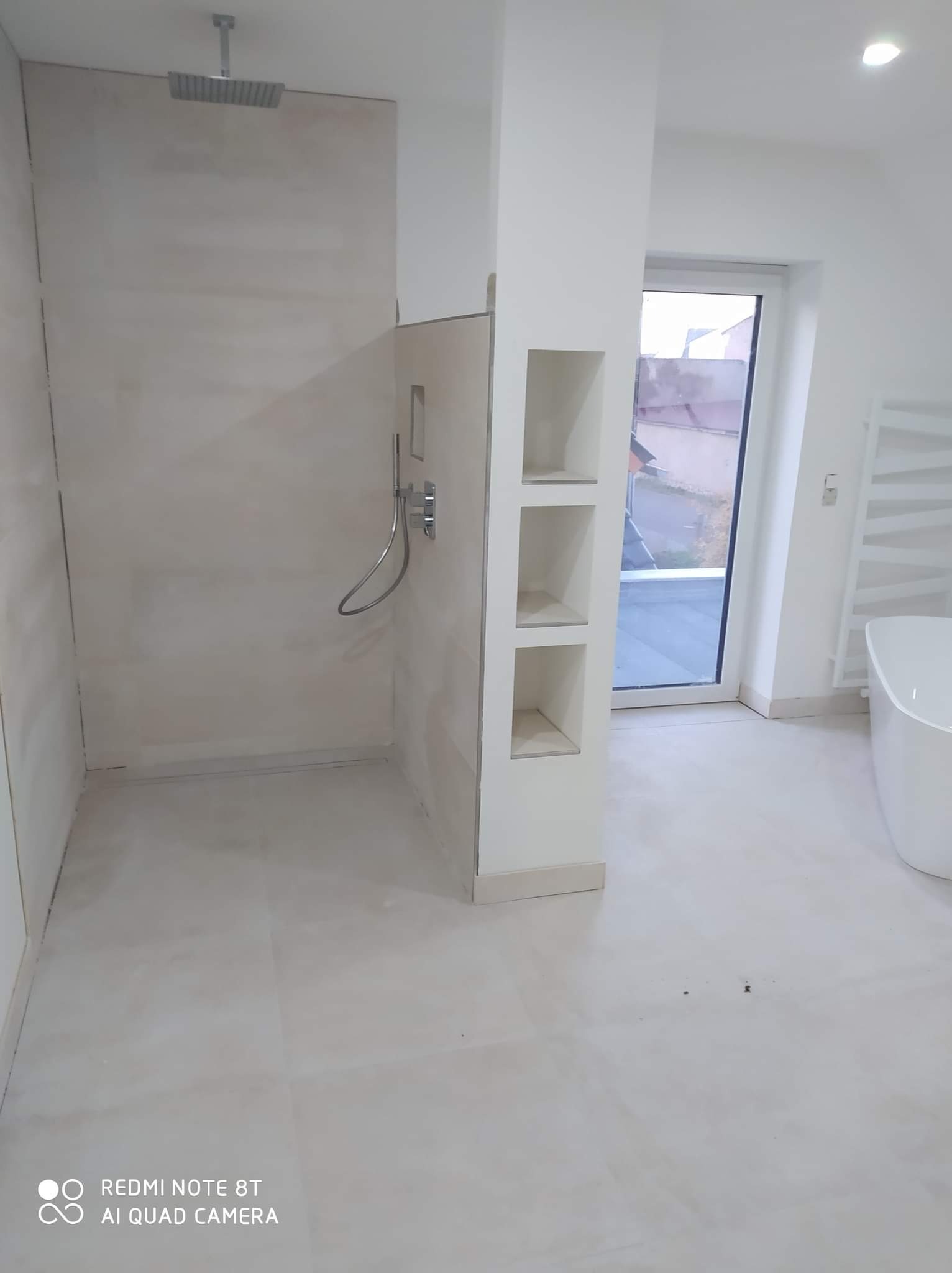 Wnętrze łazienki w trakcie prac wykończeniowych: przygotowana strefa prysznicowa z deszczownicą, fragment wanny wolnostojącej i wnęka z półkami, widoczne ślady prac na ścianach.