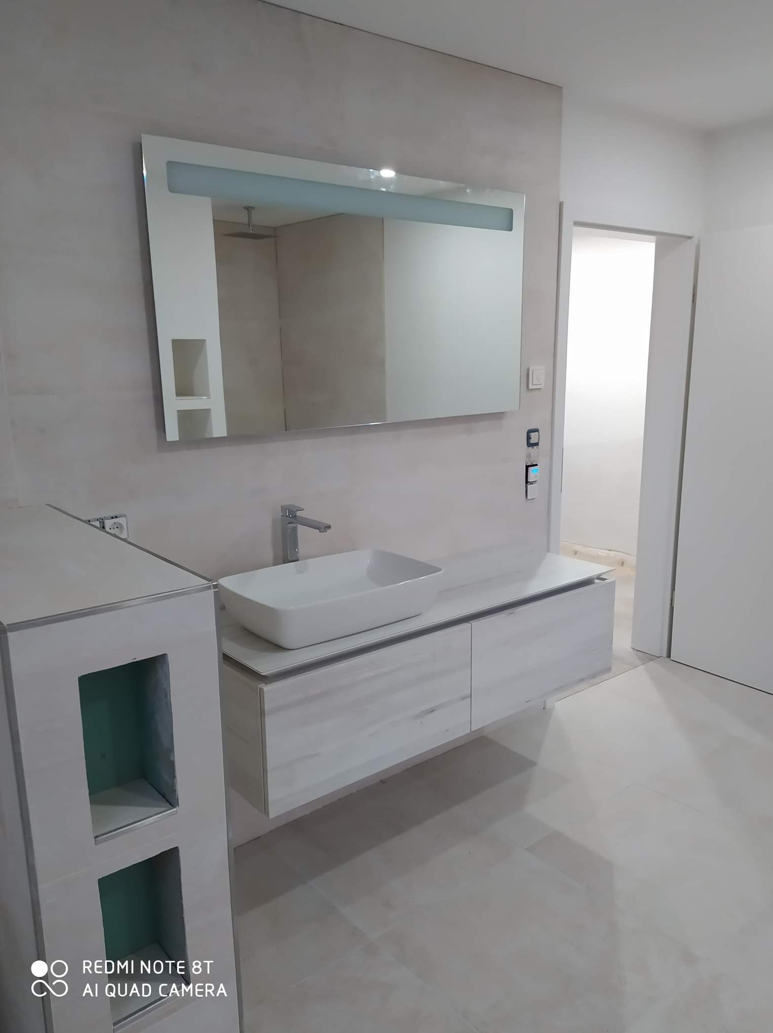 Nowoczesna łazienka z umywalką nablatową, podświetlanym lustrem i szafką z szufladami w odcieniach bieli i szarości, ściany wykończone dekoracyjnym tynkiem imitującym beton.