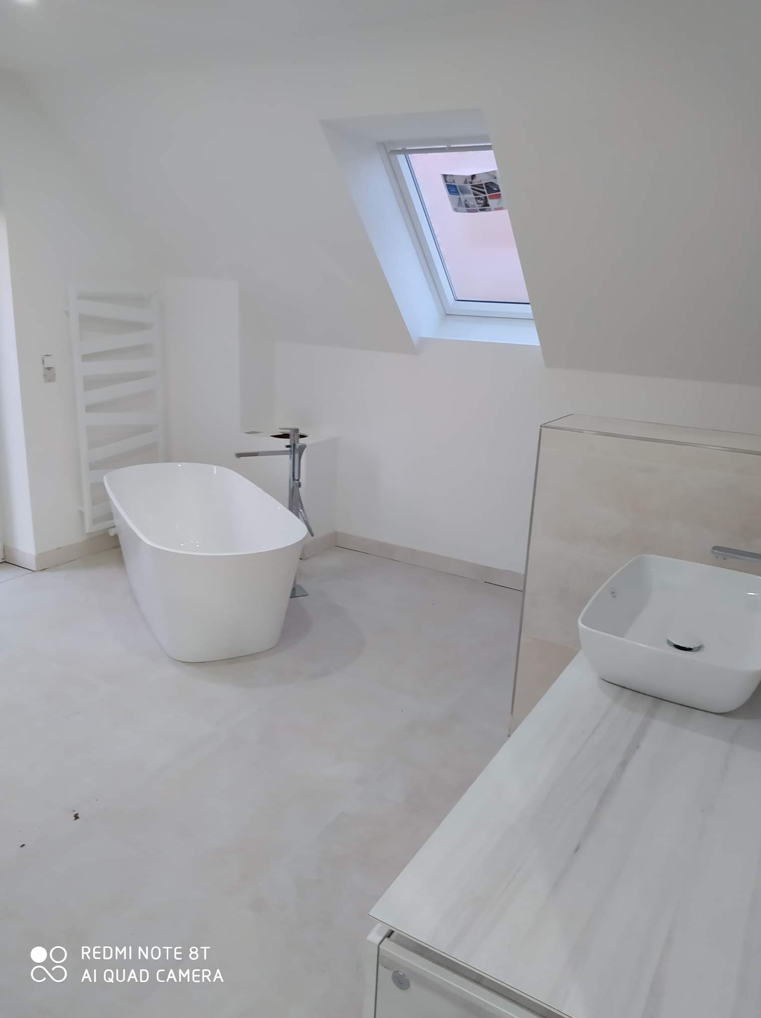 Minimalistyczna, biała łazienka na poddaszu z wolnostojącą wanną, grzejnikiem drabinkowym i umywalką nablatową, oświetlona światłem wpadającym przez okno dachowe.