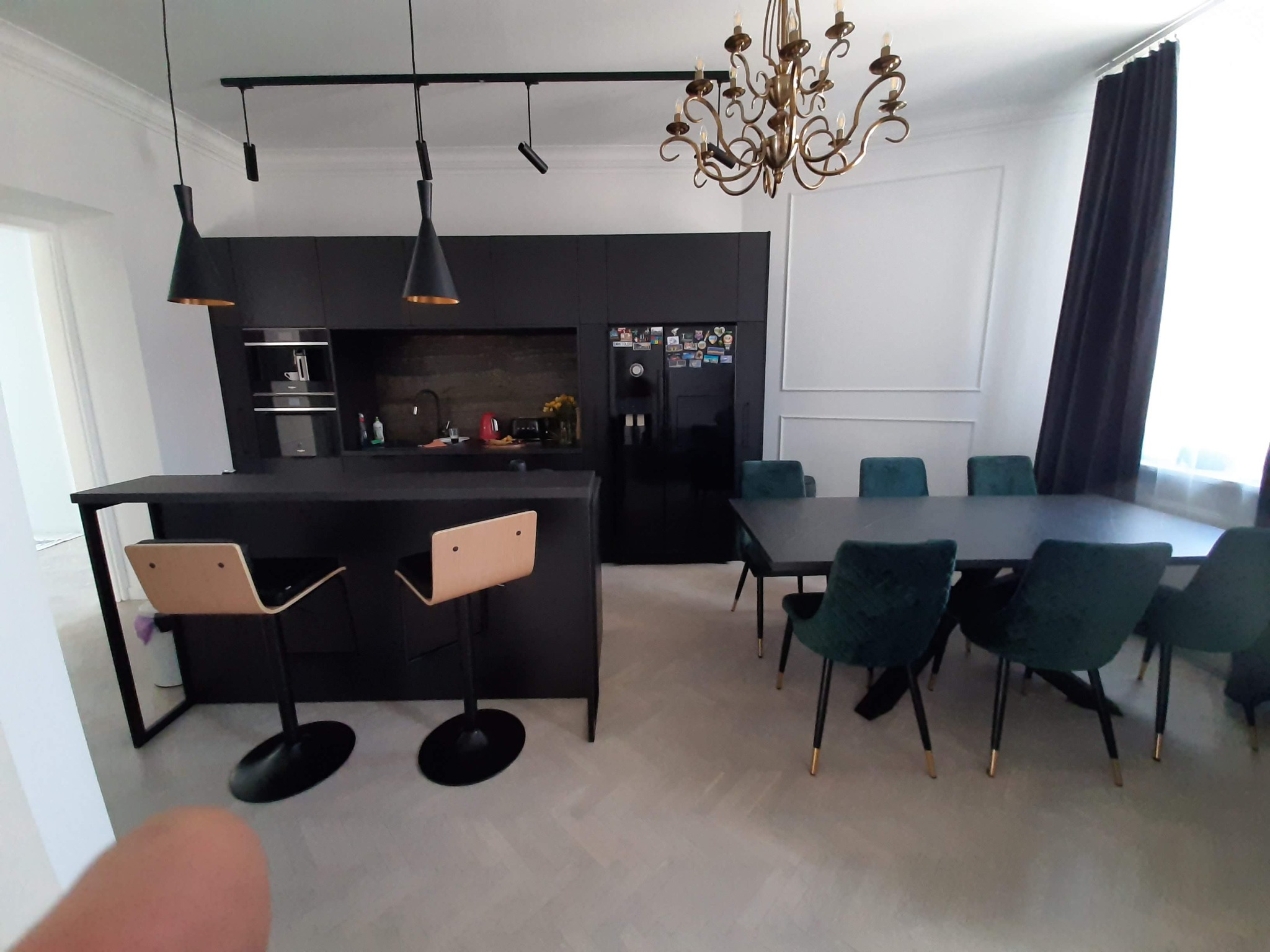 Nowoczesna kuchnia z czarnymi szafkami, wyspą z hokerami i stołem z zielonymi krzesłami, oświetlona złotym żyrandolem.