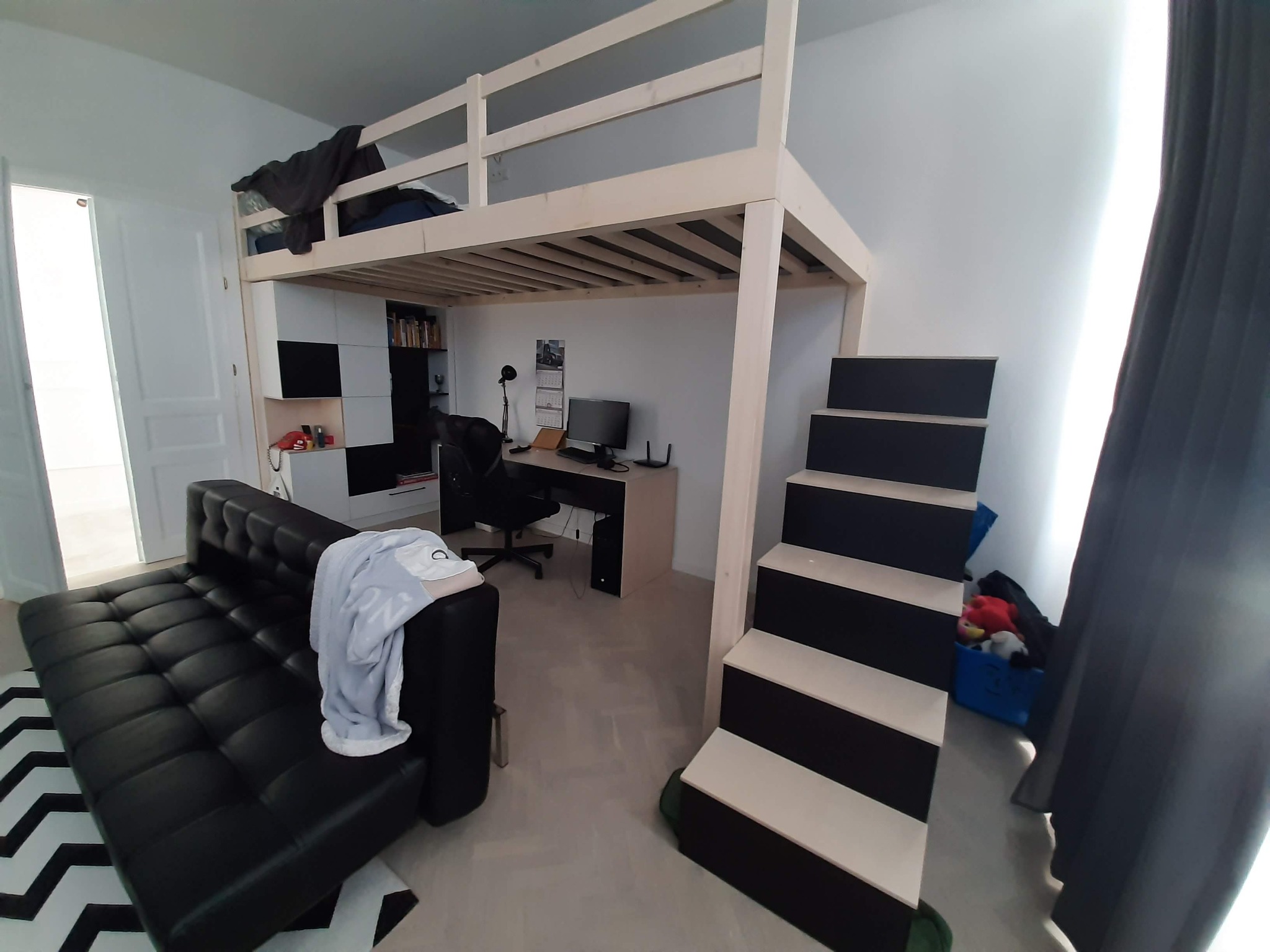 Pokój młodzieżowy z drewnianym łóżkiem na antresoli, pod którym znajduje się biurko i szafki, obok czarna sofa i schody w czarno-białe pasy.