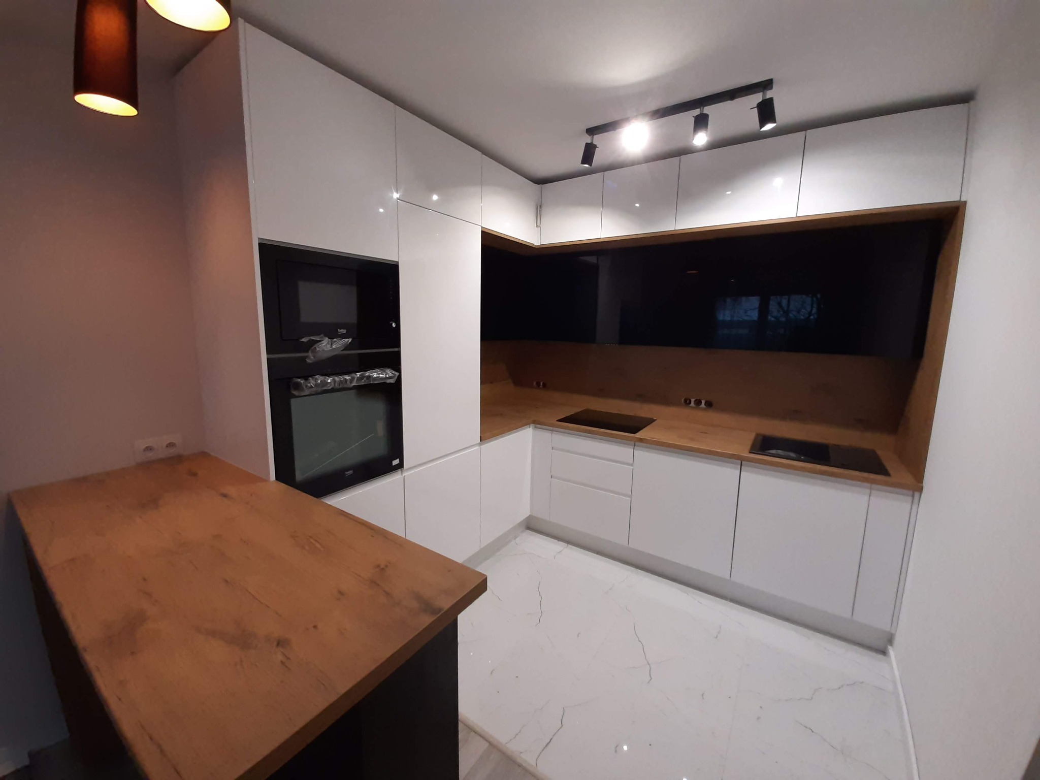 Nowoczesna kuchnia z białymi szafkami, drewnianym blatem i czarnym panelem ściennym, oświetlona lampami podszafkowymi i wiszącą lampą.