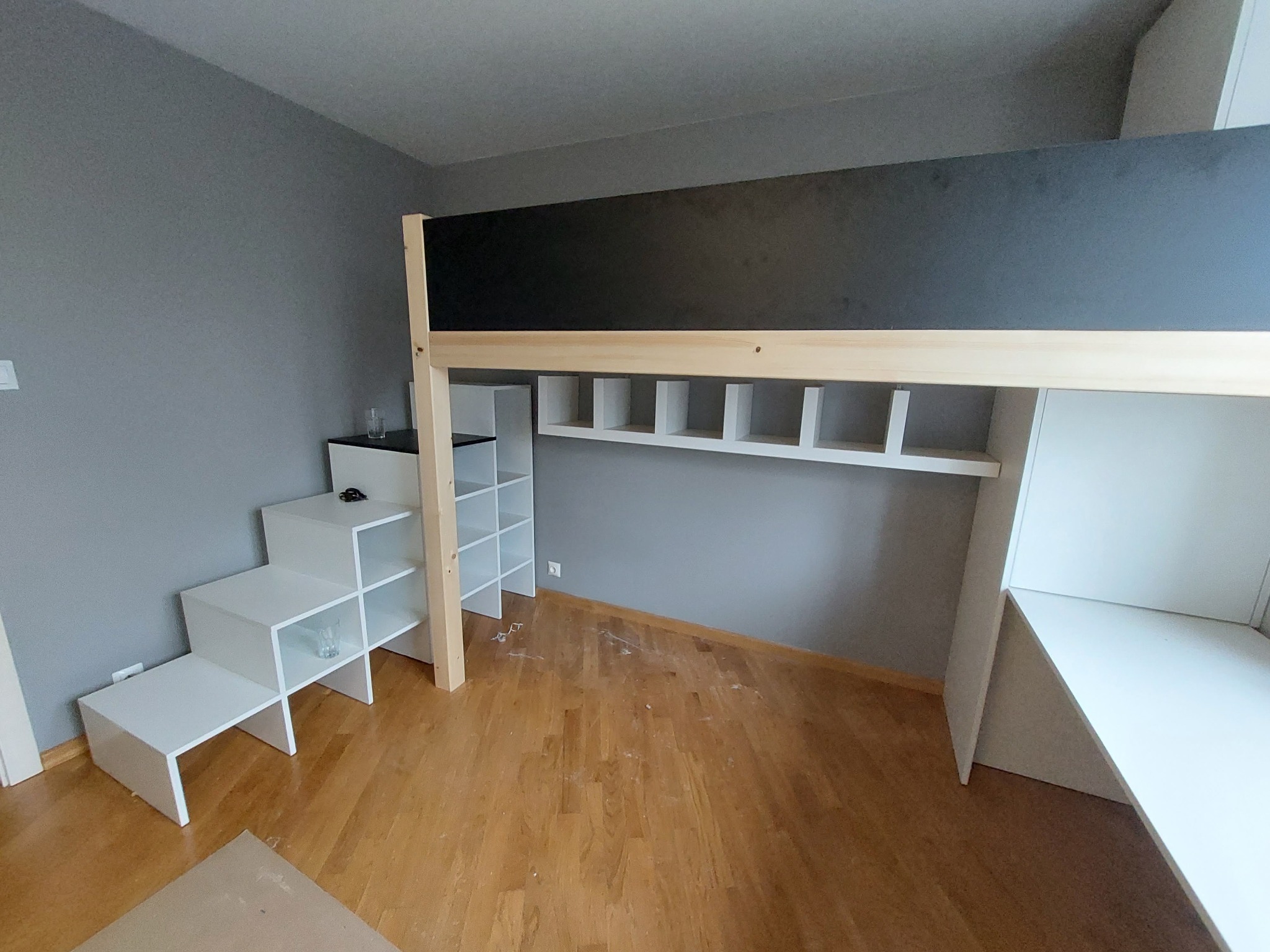Wysokie łóżko z biurkiem i schodami z szafkami w pokoju z szarą ścianą i drewnianą podłogą, widok z dołu na konstrukcję z jasnego drewna i czarną górną płytą.