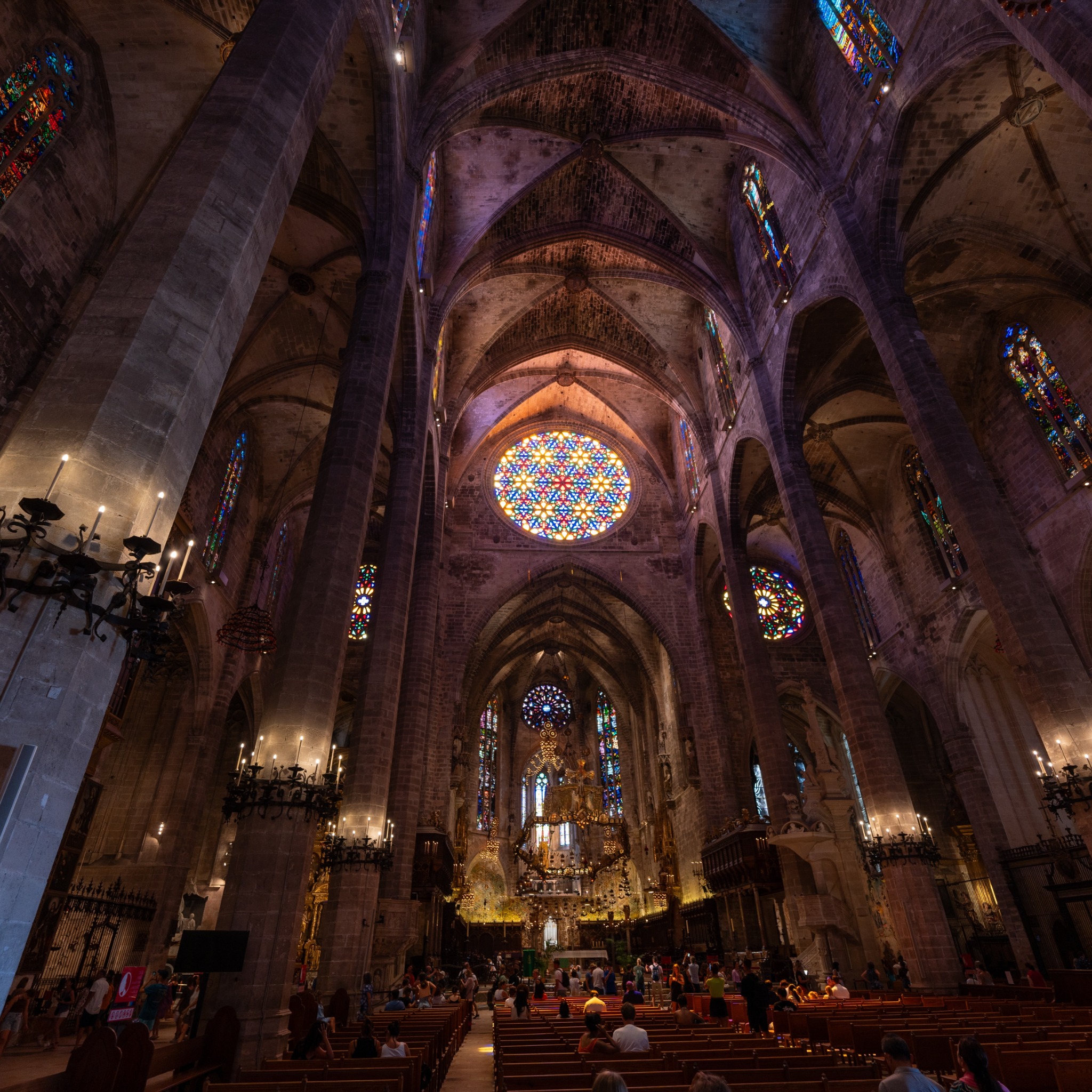 Wnętrze gotyckiej katedry z wysokimi kolumnami, witrażami i ławkami dla wiernych, oświetlone świecami i promieniami słońca wpadającymi przez okna.