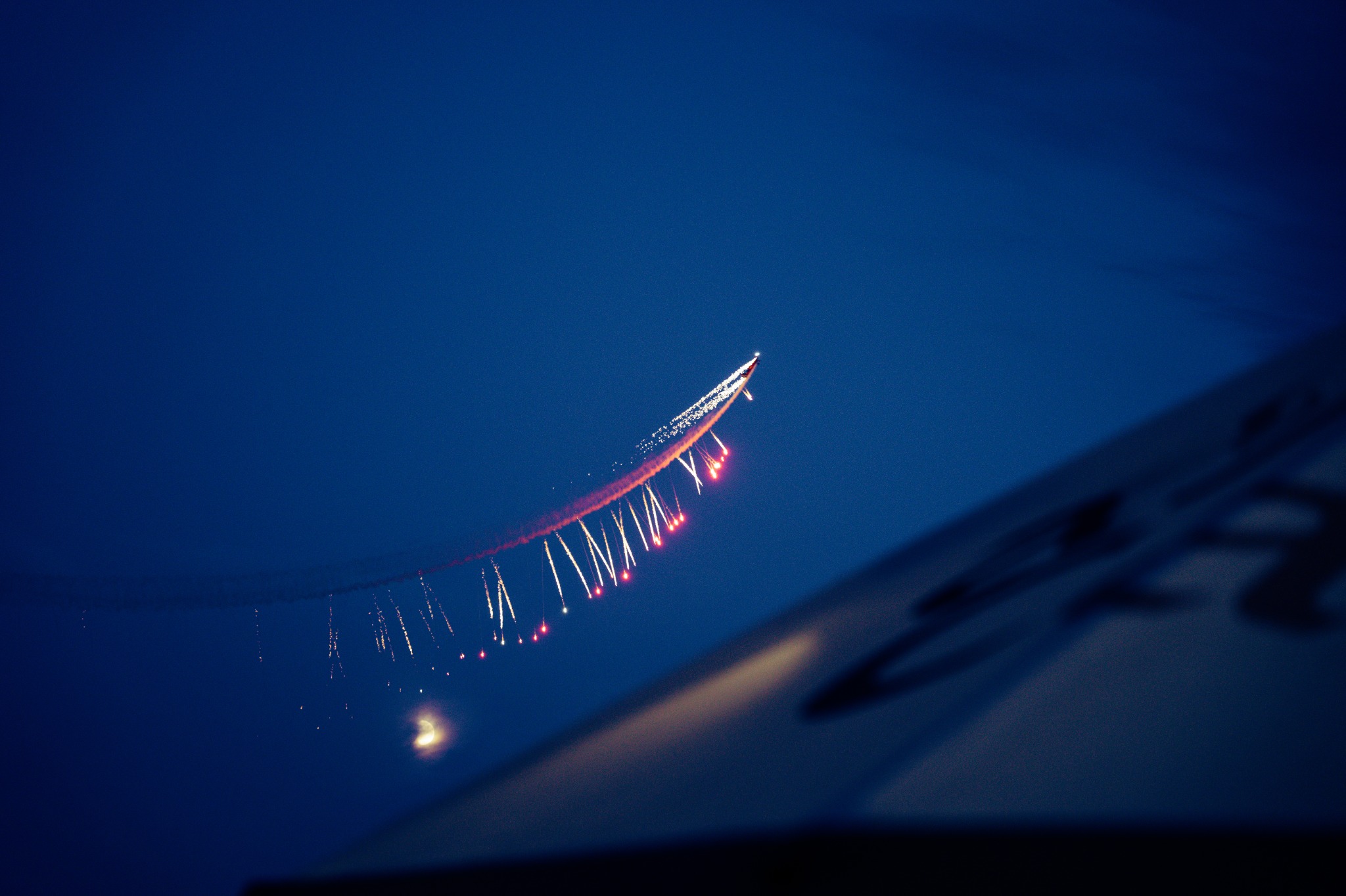Samolot zostawiający za sobą smugę z czerwonych i białych świateł na tle nocnego nieba, częściowo zasłonięty przez krawędź jasnej powierzchni w prawym dolnym rogu.