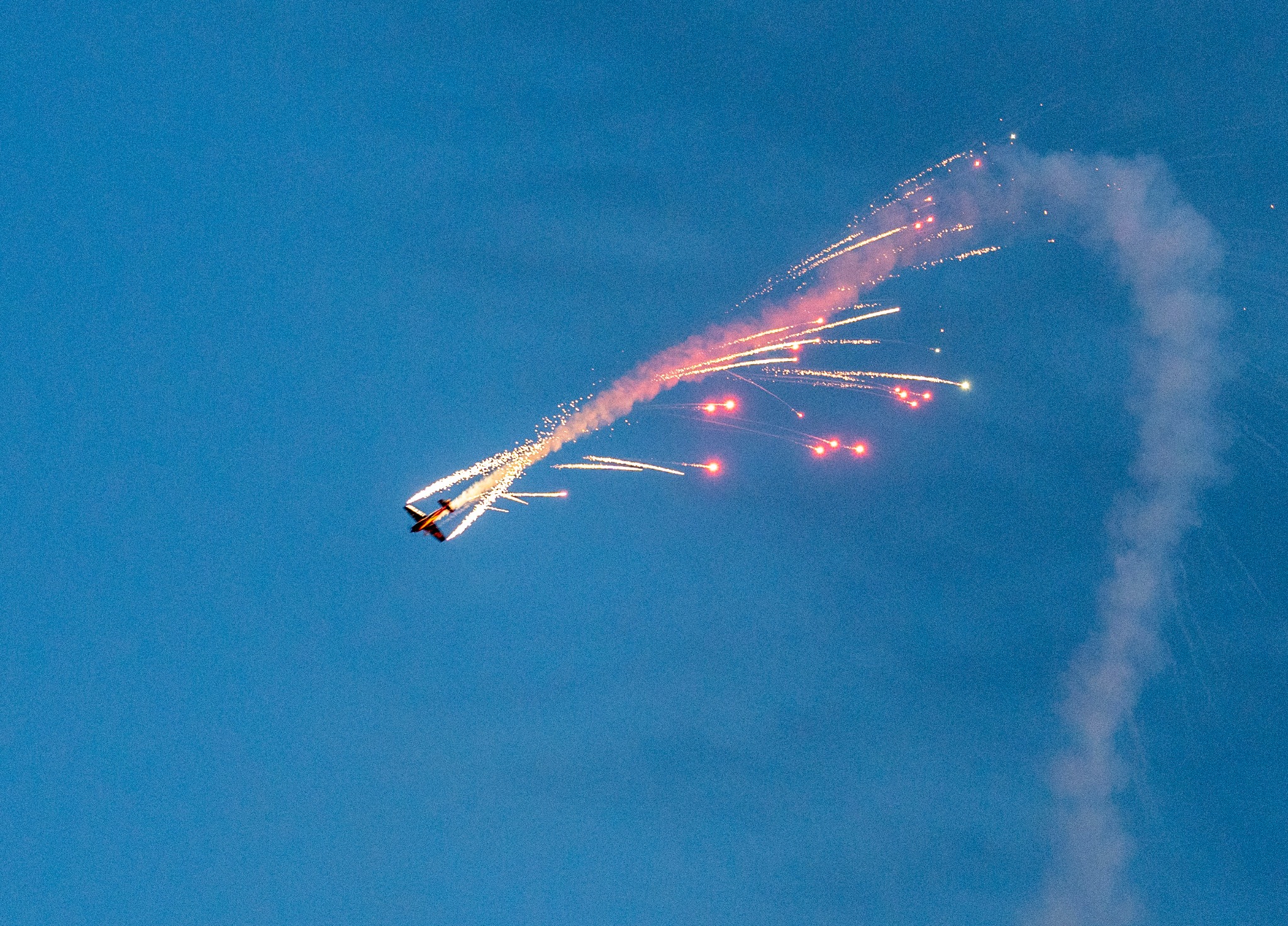 Samolot akrobacyjny na tle błękitnego nieba wykonujący pokaz z pirotechniką, pozostawiający za sobą smugę dymu i iskier.