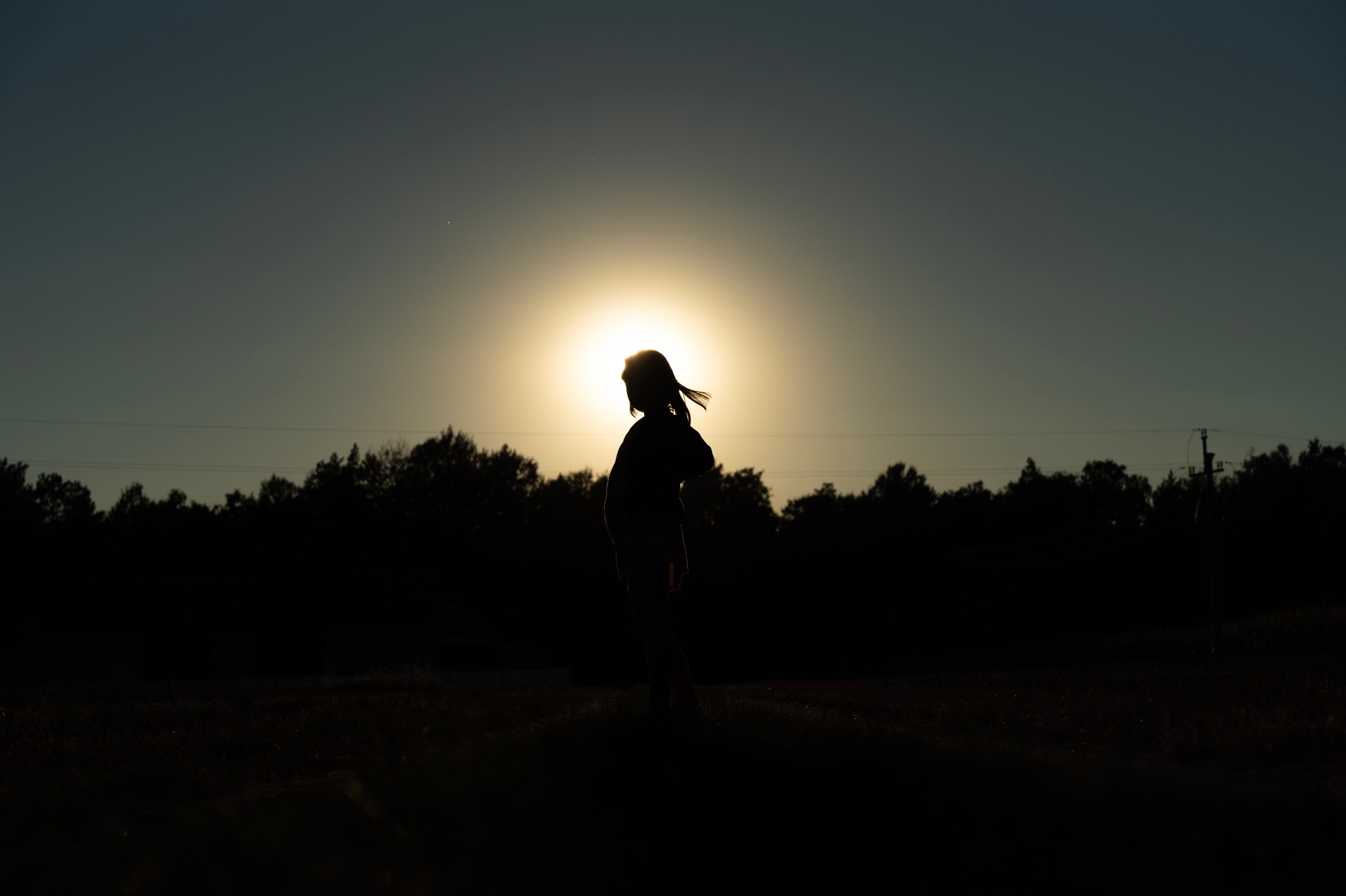 Sylwetka kobiety na tle zachodzącego słońca, włosy rozwiane, w tle ciemny pas drzew i słupy energetyczne.