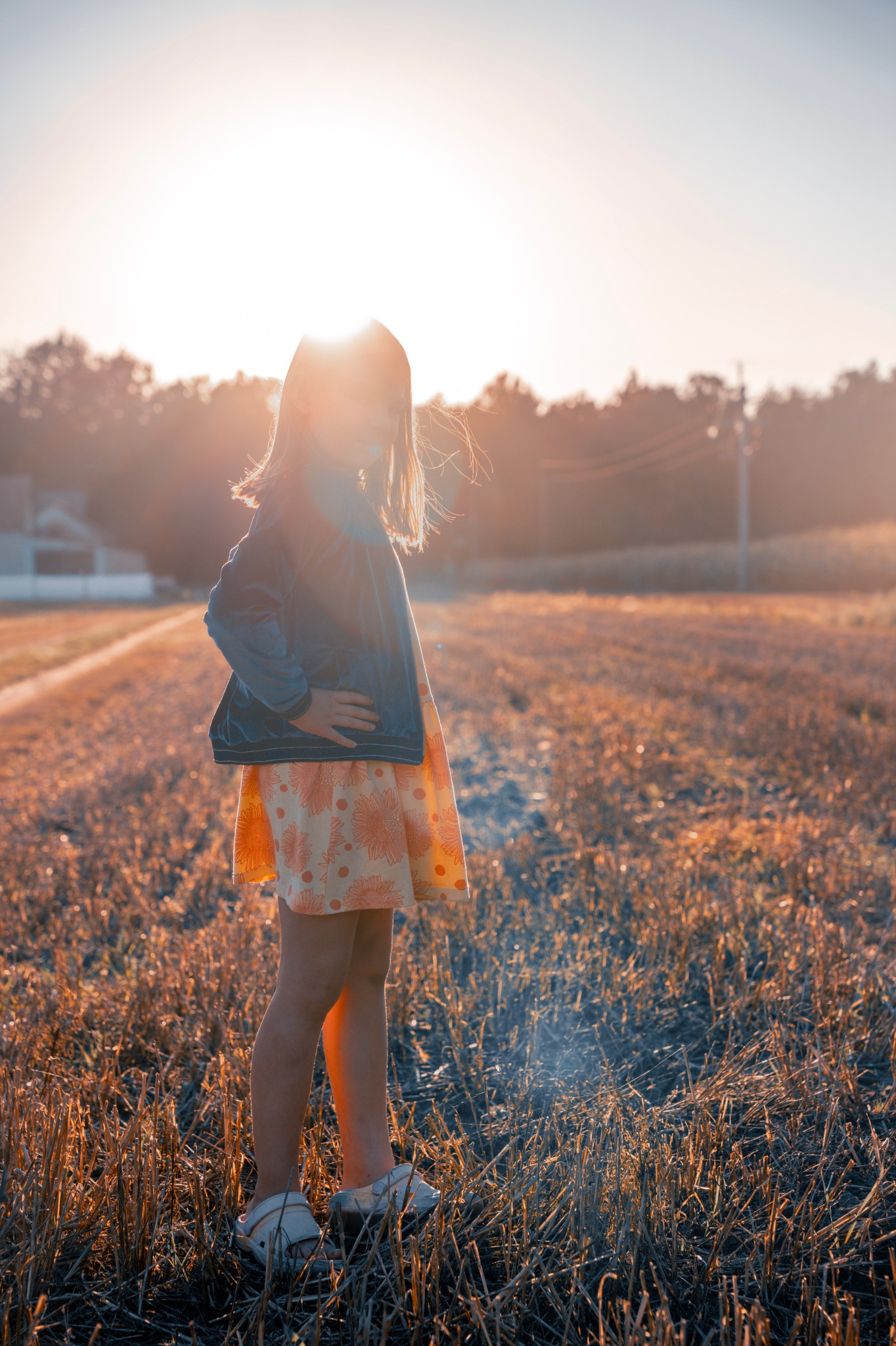 Dziewczynka w sukience z motywem słoneczników i dżinsowej kurtce stoi na polu w promieniach zachodzącego słońca, które oświetla jej sylwetkę od tyłu.