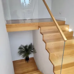 Nowoczesne schody drewniane z balustradą szklaną i drewnianym pochwytem, oświetlone punktowo, z widokiem na roślinę doniczkową na podeście.