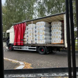 Ciężarówka z otwartą naczepą załadowana sprasowanymi rolkami materiału izolacyjnego w przezroczystej folii, widoczna przez metalowe pręty ogrodzenia, z opadłymi liśćmi na chodniku i drzewami w tle.