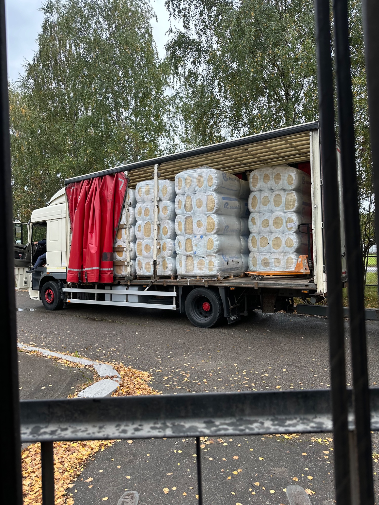 Ciężarówka z otwartą naczepą załadowana sprasowanymi rolkami materiału izolacyjnego w przezroczystej folii, widoczna przez metalowe pręty ogrodzenia, z opadłymi liśćmi na chodniku i drzewami w tle.