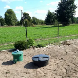 Dwa zbiorniki na piasku, jeden zielony z czerwoną pokrywką, drugi czarny z pokrywą w kształcie krzyża, otoczone siatką ogrodzeniową i zielonym polem.