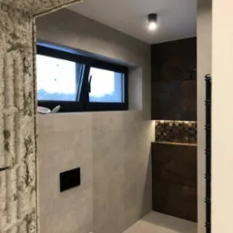 Nowoczesna łazienka z oknem, wykończona w szarościach i brązach, widoczna przez surowy otwór drzwiowy z odsłoniętym betonem, minimalistyczna toaleta pod oknem.