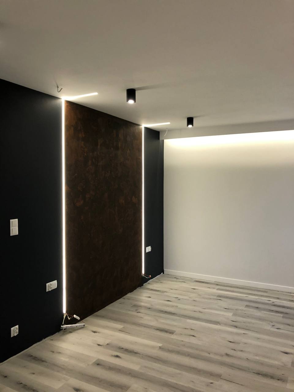 Nowoczesne wnętrze z oświetleniem LED w pionowych listwach, ciemna ściana z dekoracyjną fakturą, jasna podłoga imitująca drewno, minimalistyczne lampy sufitowe.