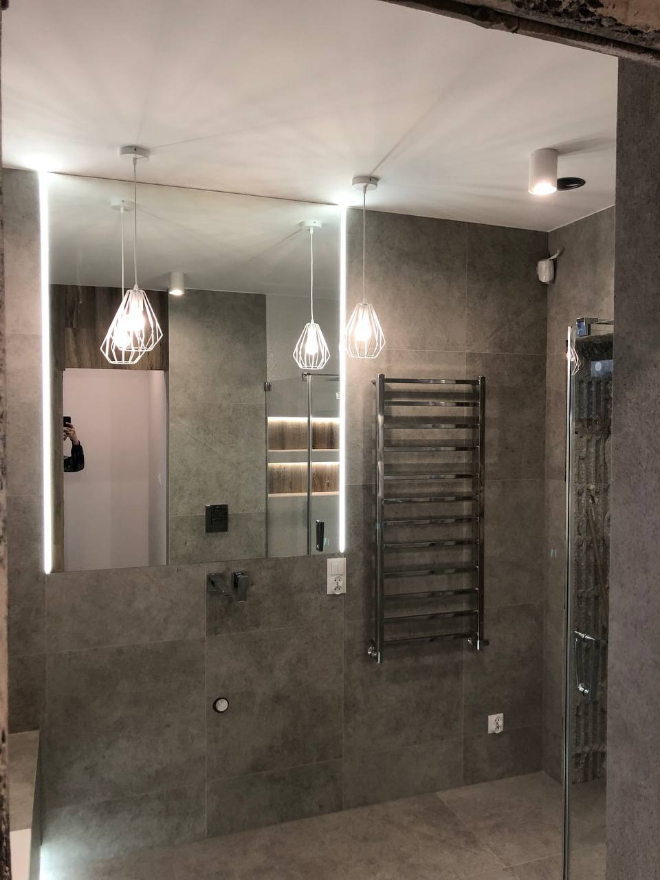 Nowoczesna łazienka z szarymi płytkami, podświetlanym lustrem, grzejnikiem drabinkowym i wiszącymi lampami z ażurowymi kloszami.