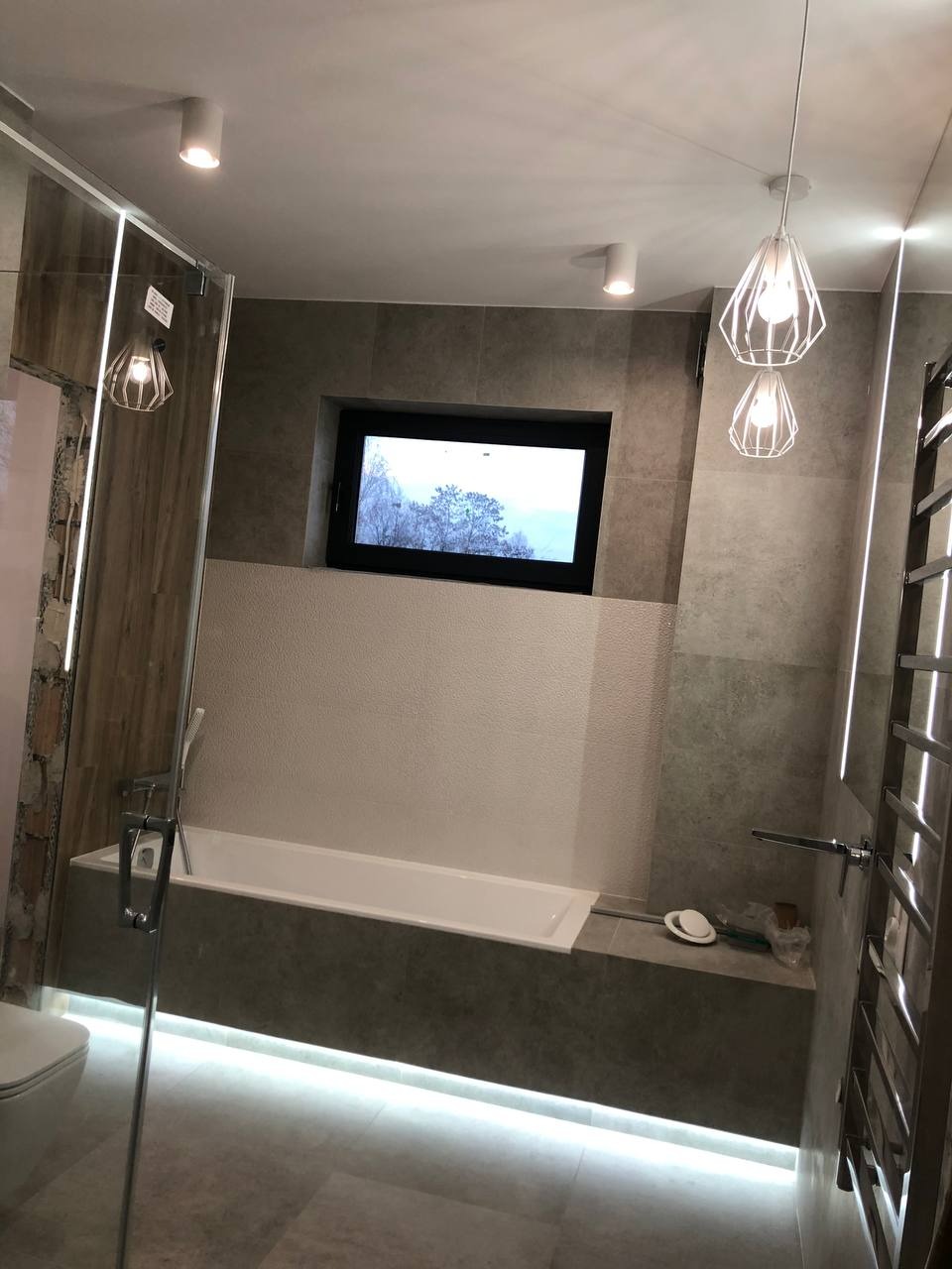 Nowoczesna łazienka z wanną pod oknem, oświetleniem LED pod wanną i dekoracyjnymi lampami wiszącymi, wykończona w szarościach i bieli.