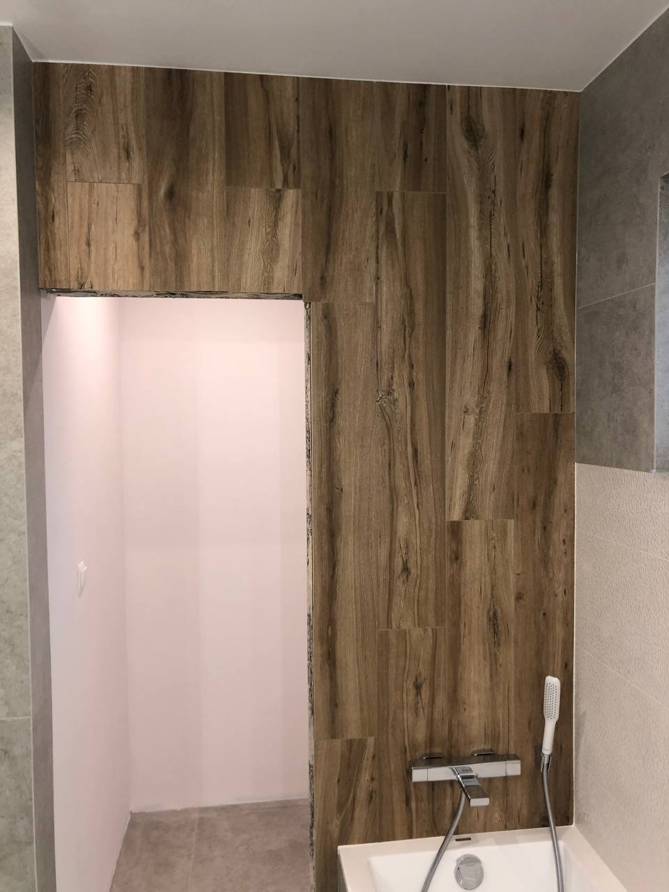 Nowoczesna łazienka z obudową wanny i ściany wyłożoną płytkami imitującymi drewno, widoczna armatura i słuchawka prysznicowa.