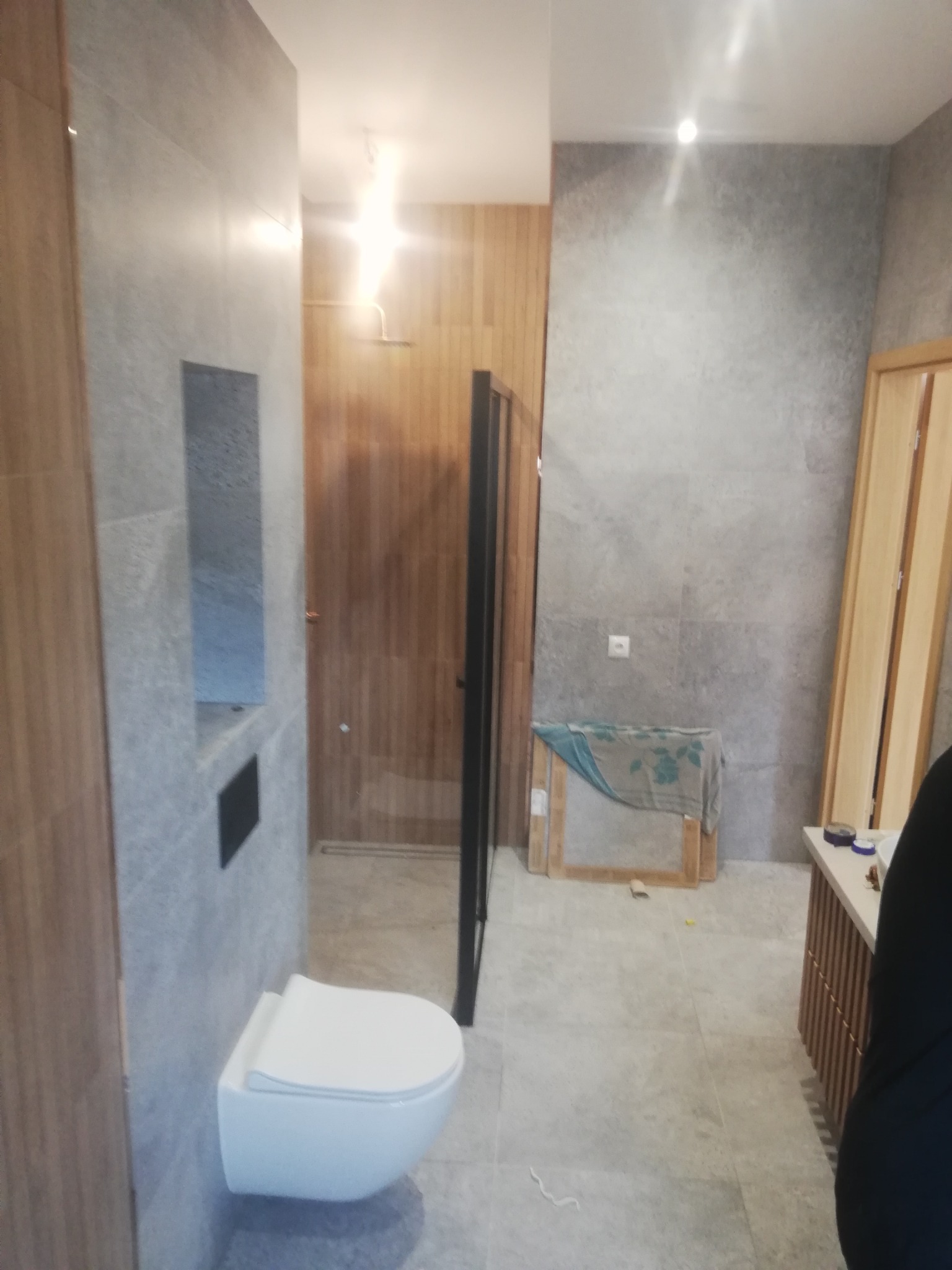 Nowoczesna łazienka w trakcie prac wykończeniowych, widoczna podwieszana toaleta, szare płytki na ścianach i podłodze, drewniane akcenty oraz niezabezpieczone drzwi po prawej stronie.