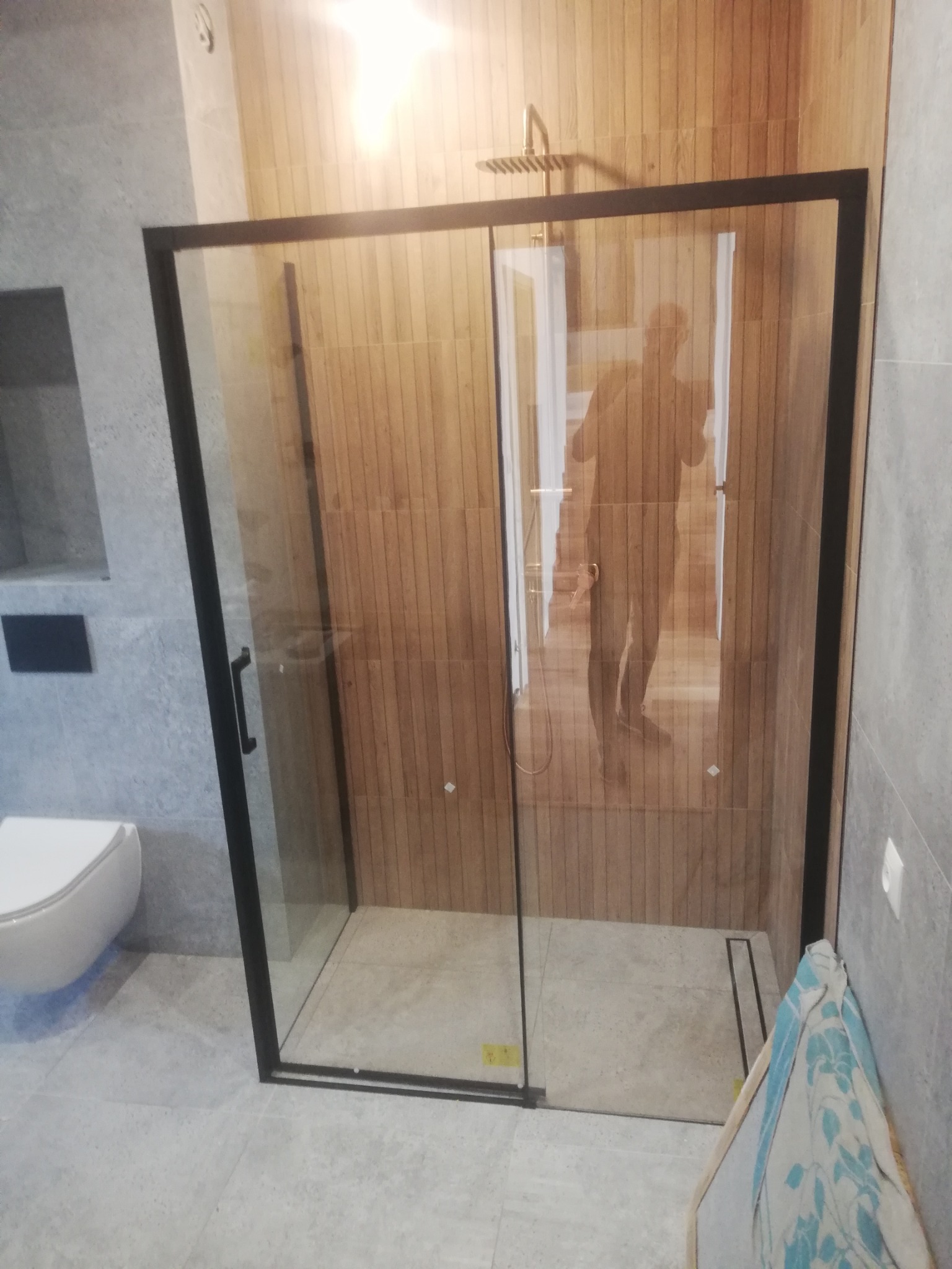 Nowoczesna kabina prysznicowa z czarną ramą i przesuwnymi drzwiami, drewnopodobne płytki na ścianach, widoczna deszczownica i odpływ liniowy, szare płytki podłogowe, fragment toalety podwieszanej.