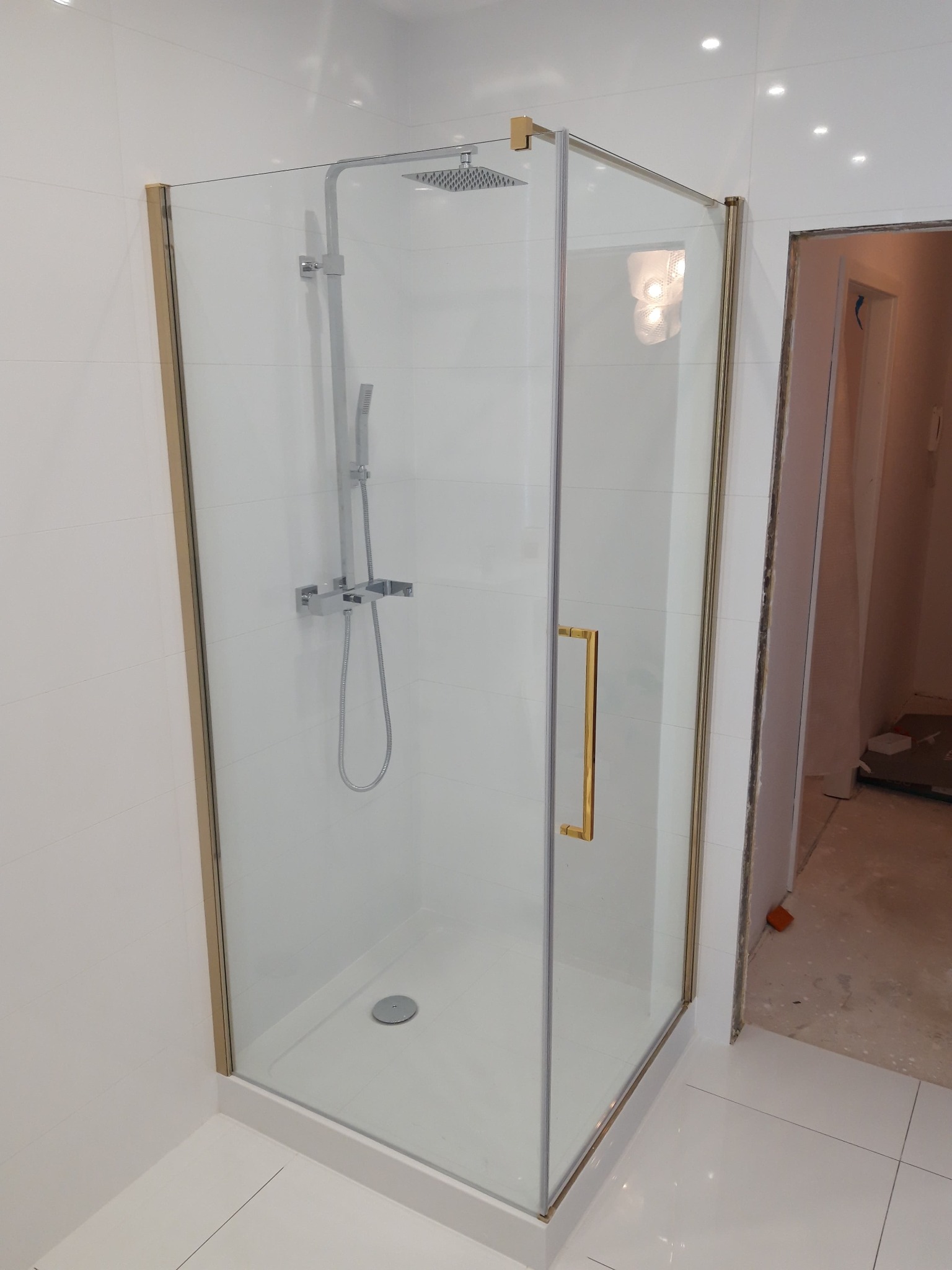 Nowoczesna kabina prysznicowa z chromowaną armaturą i złotymi profilami, widoczna na tle białych płytek w łazience w trakcie remontu.