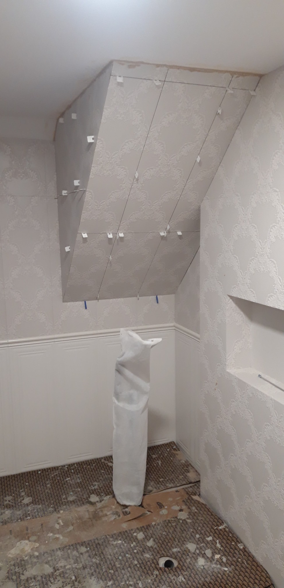 Montaż płytek na skośnym suficie z użyciem krzyżyków dystansowych i sznurków poziomujących, widoczny worek z klejem i przygotowane podłoże na podłodze.