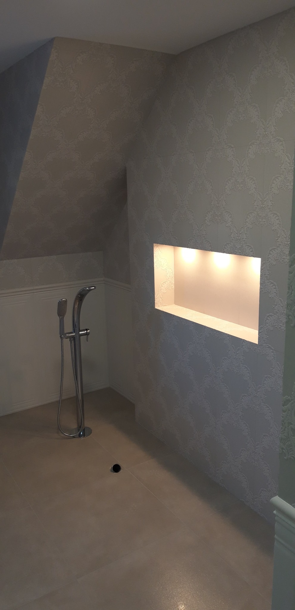 Nowoczesna łazienka w odcieniach szarości i bieli z wolnostojącą baterią prysznicową i wnęką z oświetleniem punktowym w ścianie pokrytej wzorzystymi płytkami.