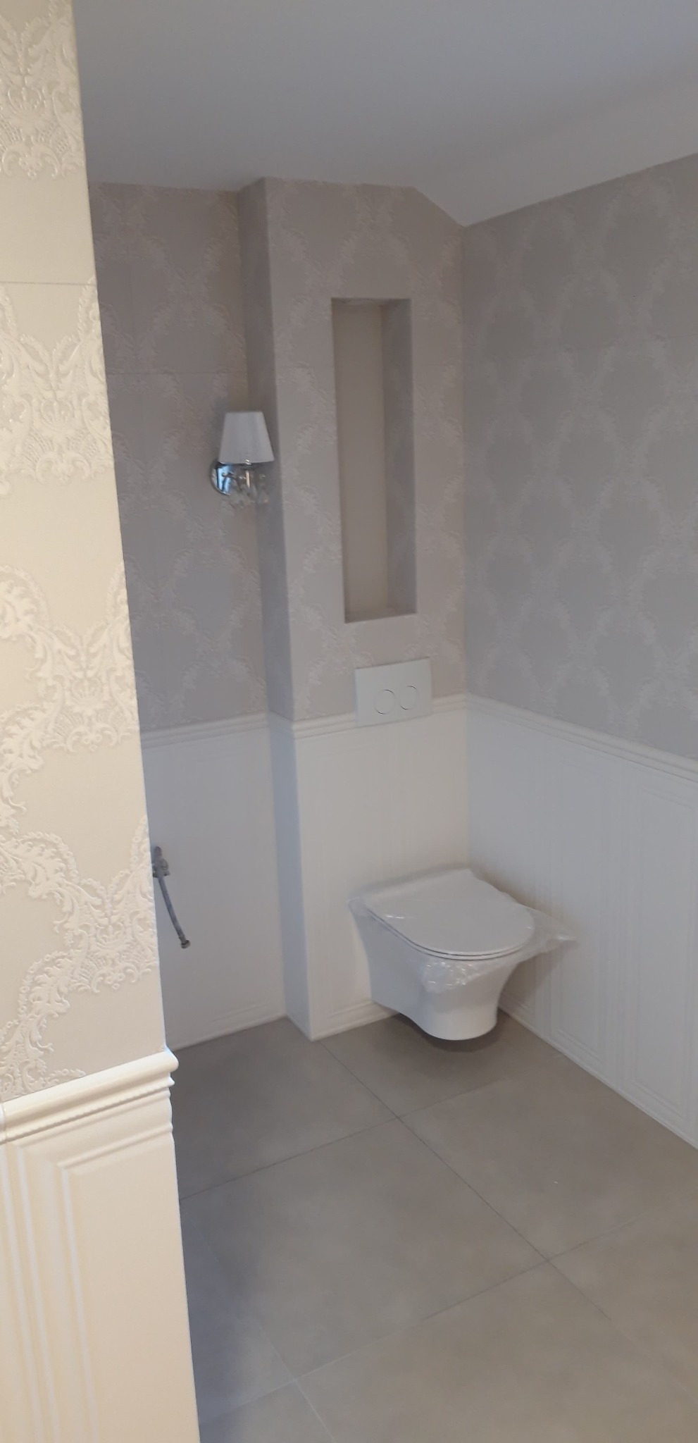 Wykończona łazienka z podwieszaną toaletą, szarą płytką podłogową, dekoracyjną tapetą w górnej części ścian i białymi panelami ściennymi w dolnej części, oświetleniem ściennym i wnęką w ścianie.