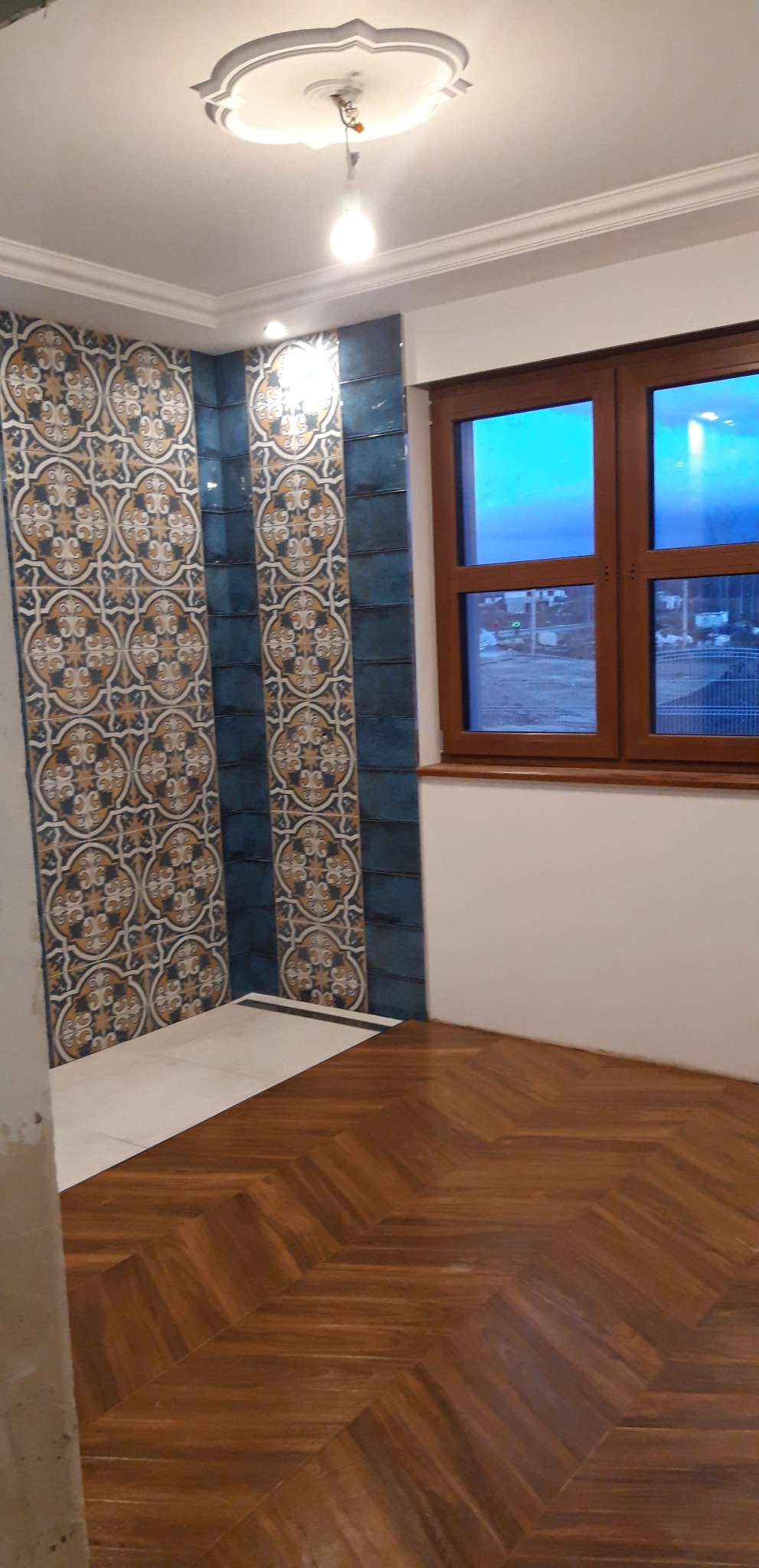 Wykończona łazienka z wzorzystymi, niebiesko-złotymi płytkami na ścianie, białym brodzikiem i drewnianą podłogą ułożoną we wzór jodełki, widok na drewniane okno.